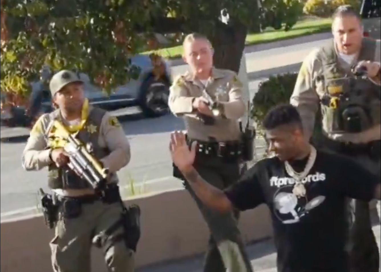 Blueface, with raised hands, is approached by three armed officers on a street. Trees and a car are visible in the background.