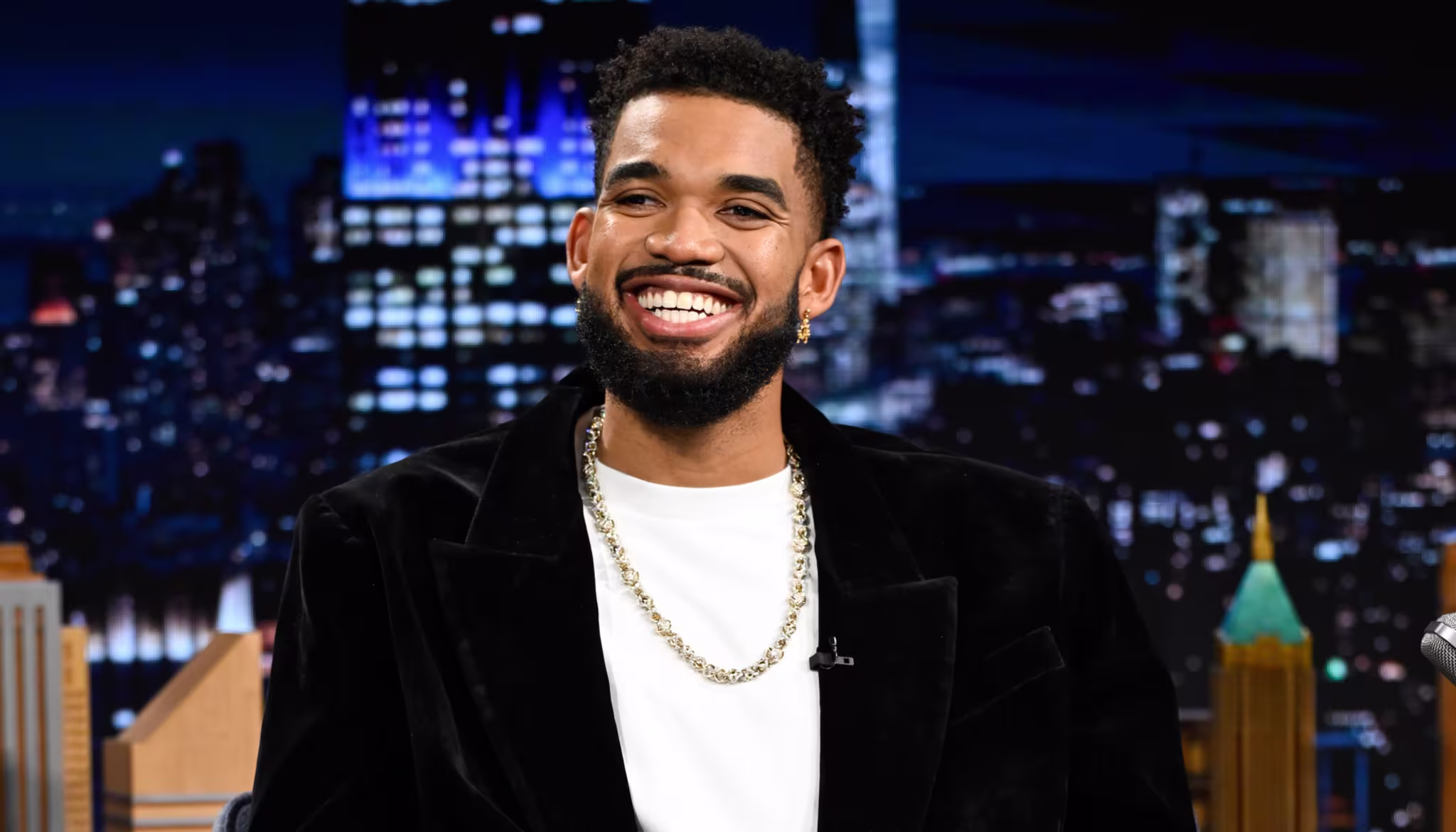 Karl-Anthony Towns in a black suit and white shirt sits on a talk show set, smiling. The background features a night cityscape.
