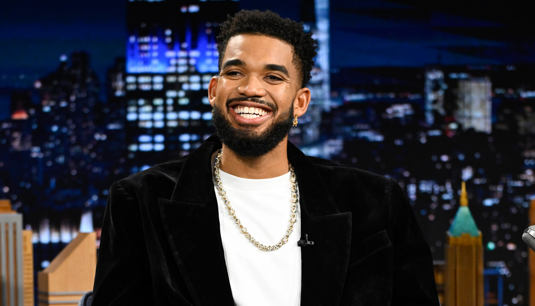 Karl-Anthony Towns in a black suit and white shirt sits on a talk show set, smiling. The background features a night cityscape.