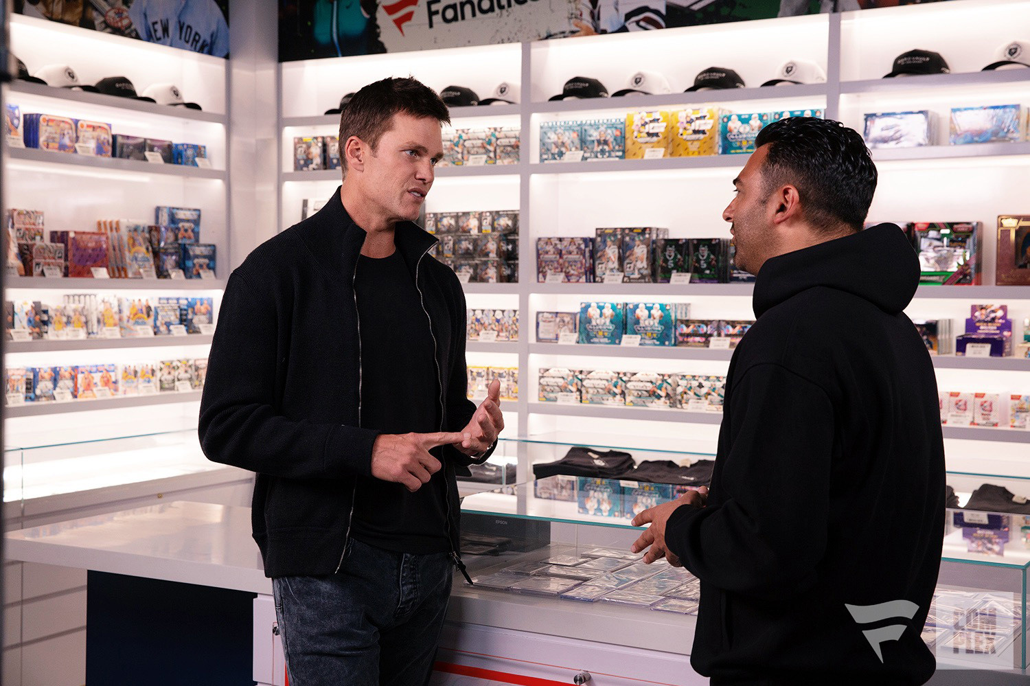 Tom Brady and 'Card Shopping' host Joe La Puma are talking in a store with sports memorabilia and collectibles on display.