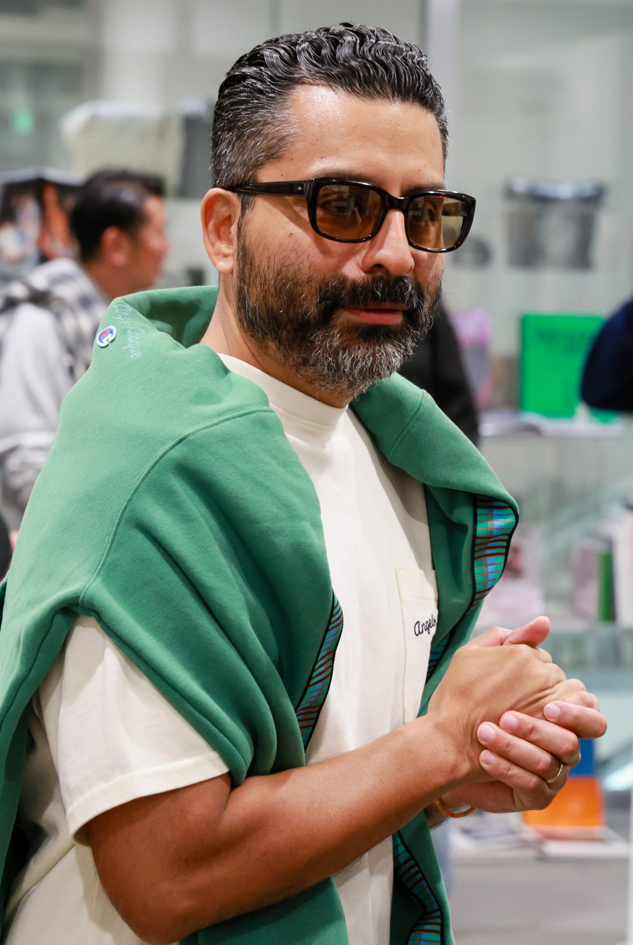 A man with a beard and sunglasses, wearing a white shirt and green sweater draped over his shoulders, stands indoors.