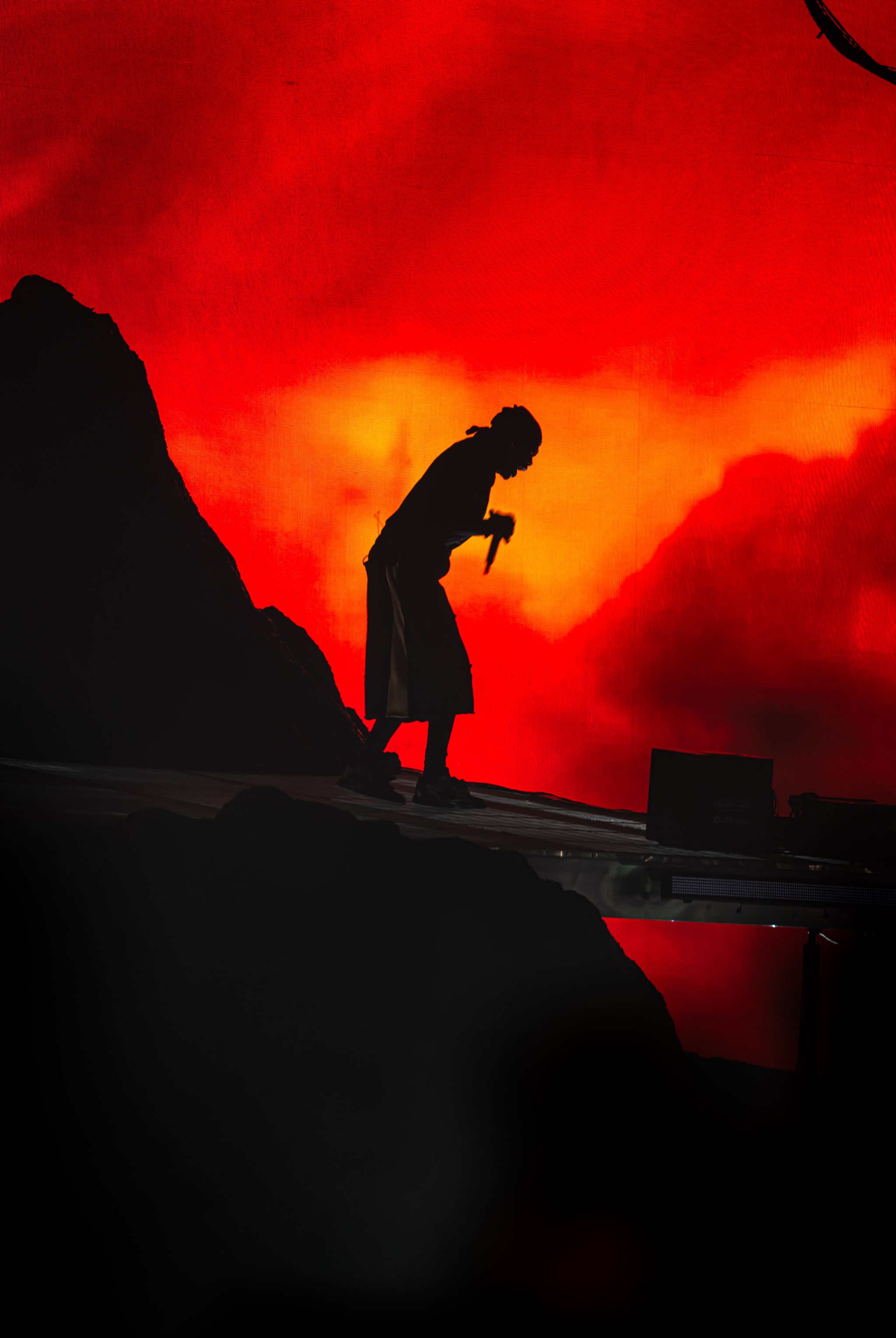 Silhouette of Travis Scott on stage with a fiery red and orange background, holding a microphone.