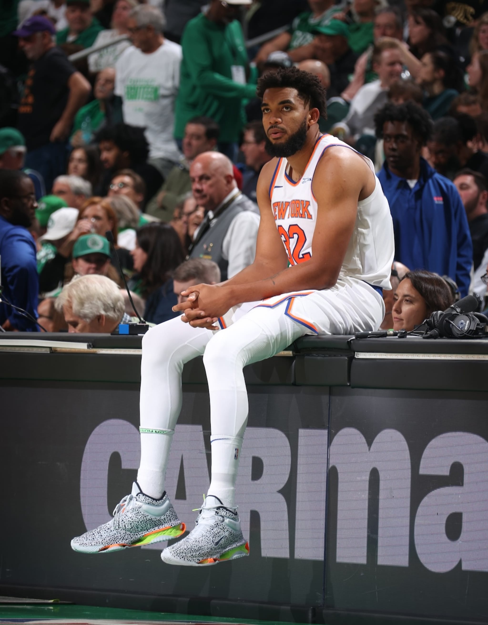 Karl Anthony-Towns in a New York jersey sits on the sideline, surrounded by a crowd. He's wearing gray sneakers with colorful accents.