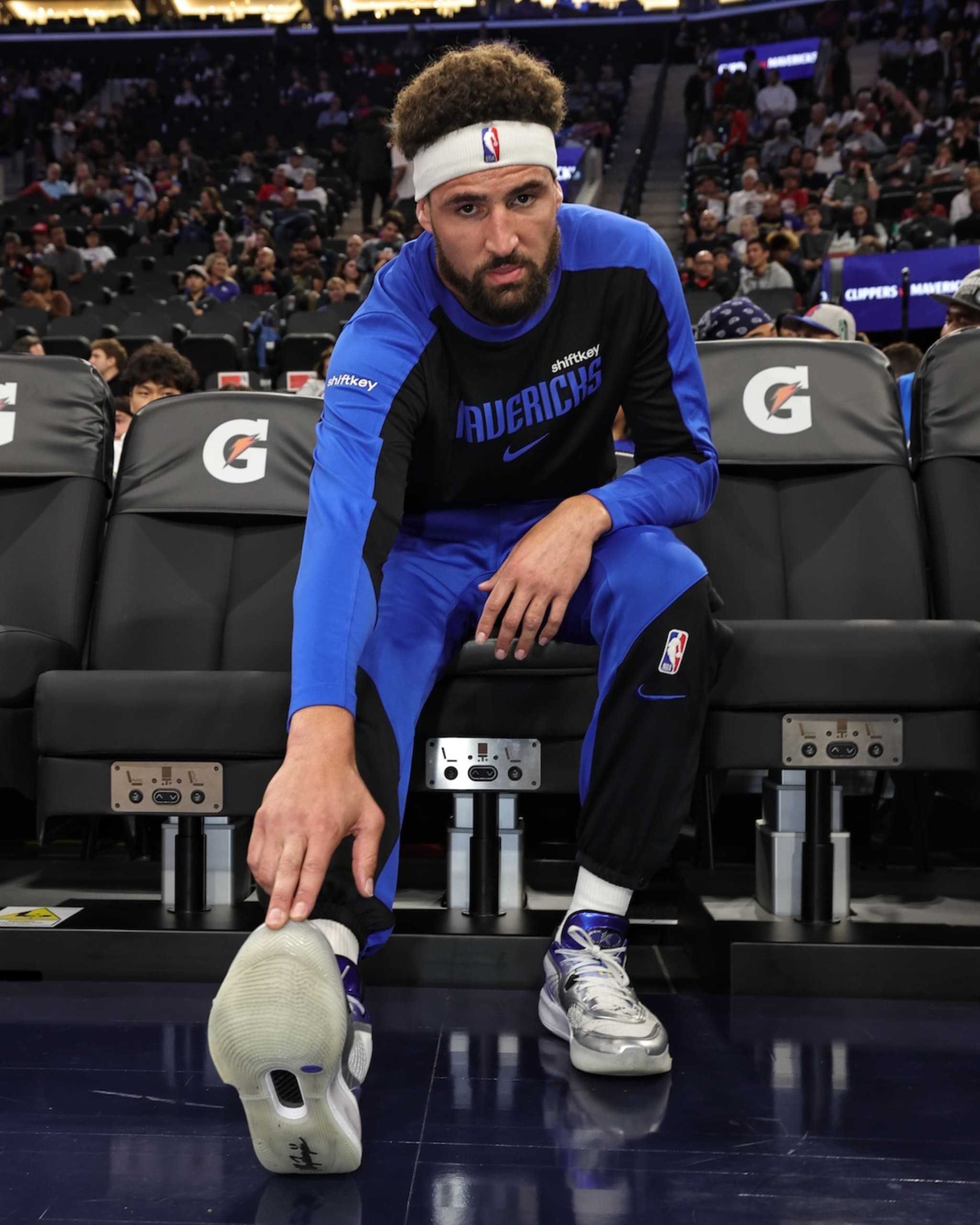 Klay Thompson in a Mavericks uniform stretches on the court, wearing a headband and sneakers, with an audience in the background.