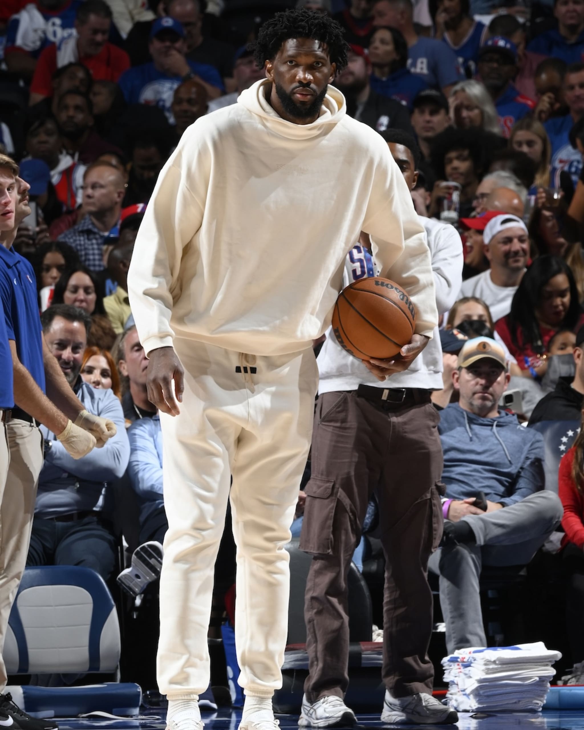 Joel Embiid, in a cream tracksuit, holds a basketball courtside at a game, with a crowd in the background.