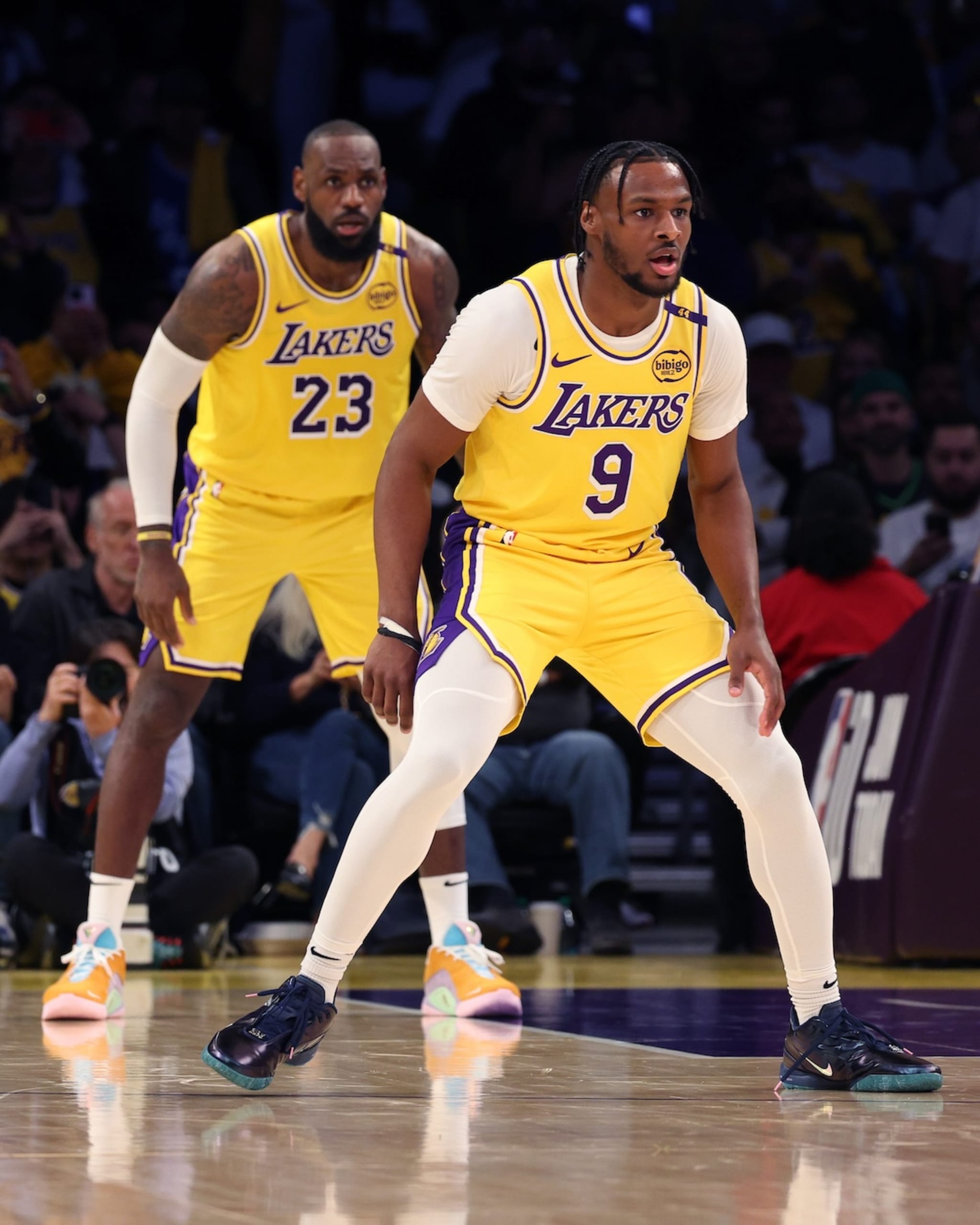 LeBron James and Bronny James in Lakers uniforms on the basketball court, focused and ready for action.
