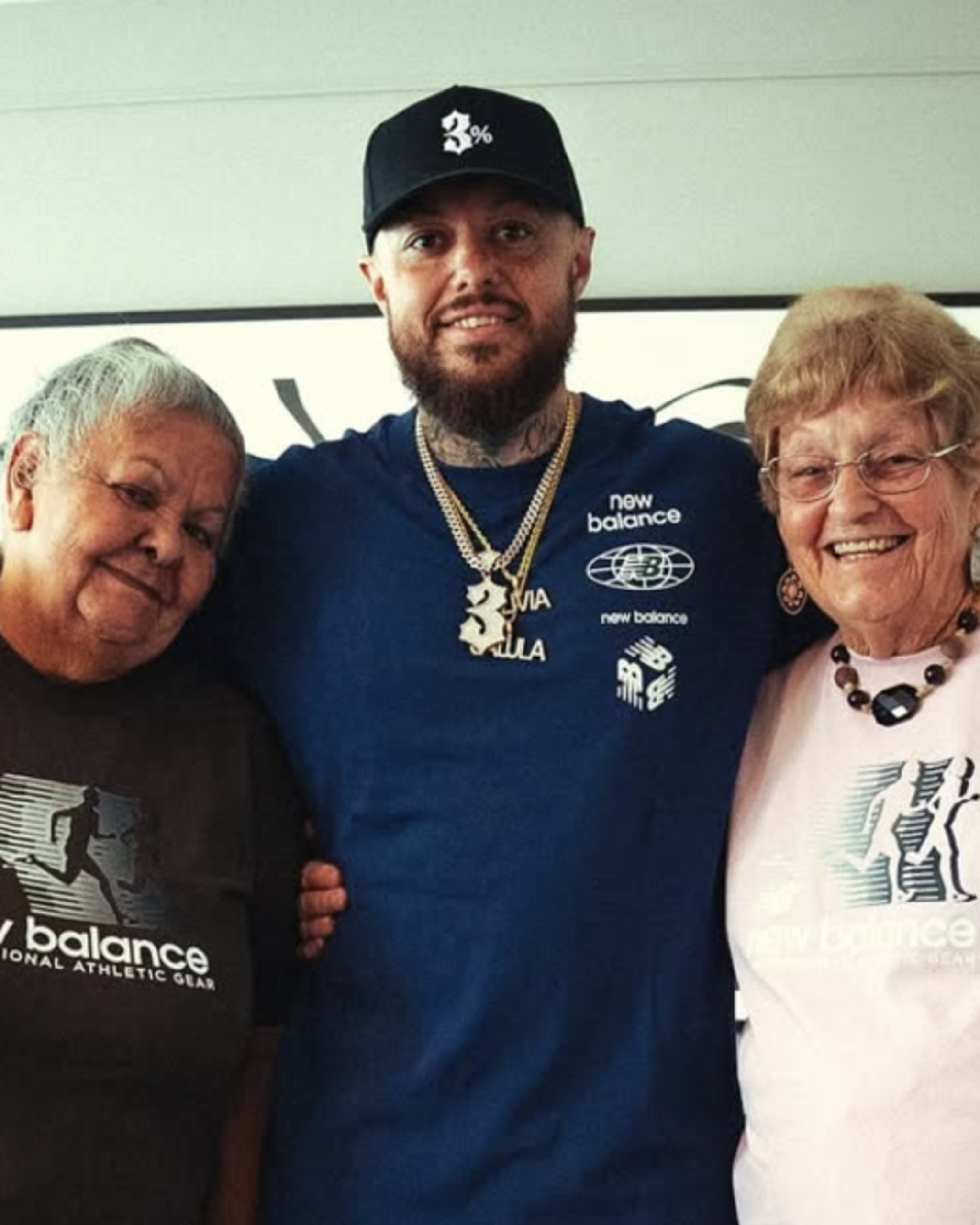 A man in a blue New Balance shirt and cap stands between two older women, each wearing New Balance shirts, smiling indoors.