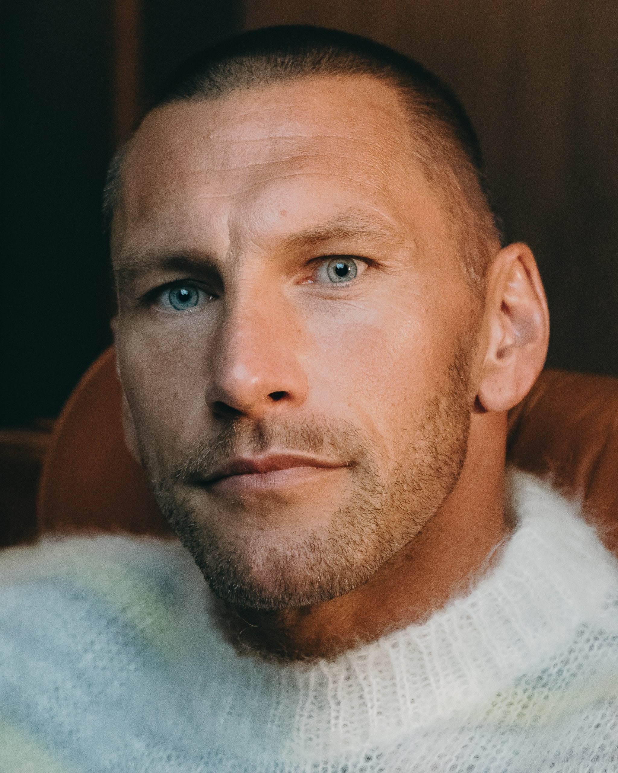 A man with short hair and a beard, wearing a fuzzy sweater, sits on a leather chair in a warmly lit room.