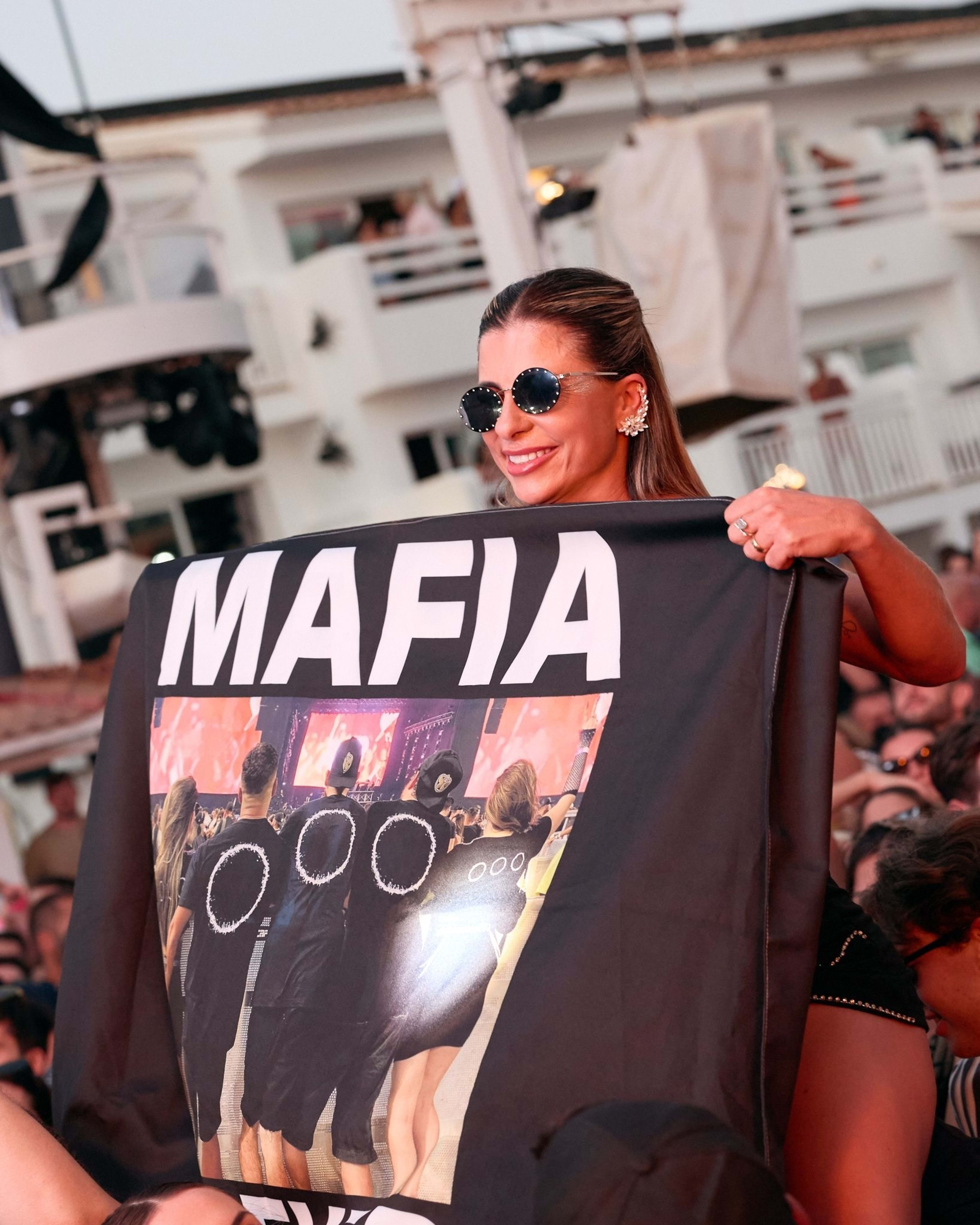A woman holds a "MAFIA" banner at an outdoor music event, smiling and wearing sunglasses