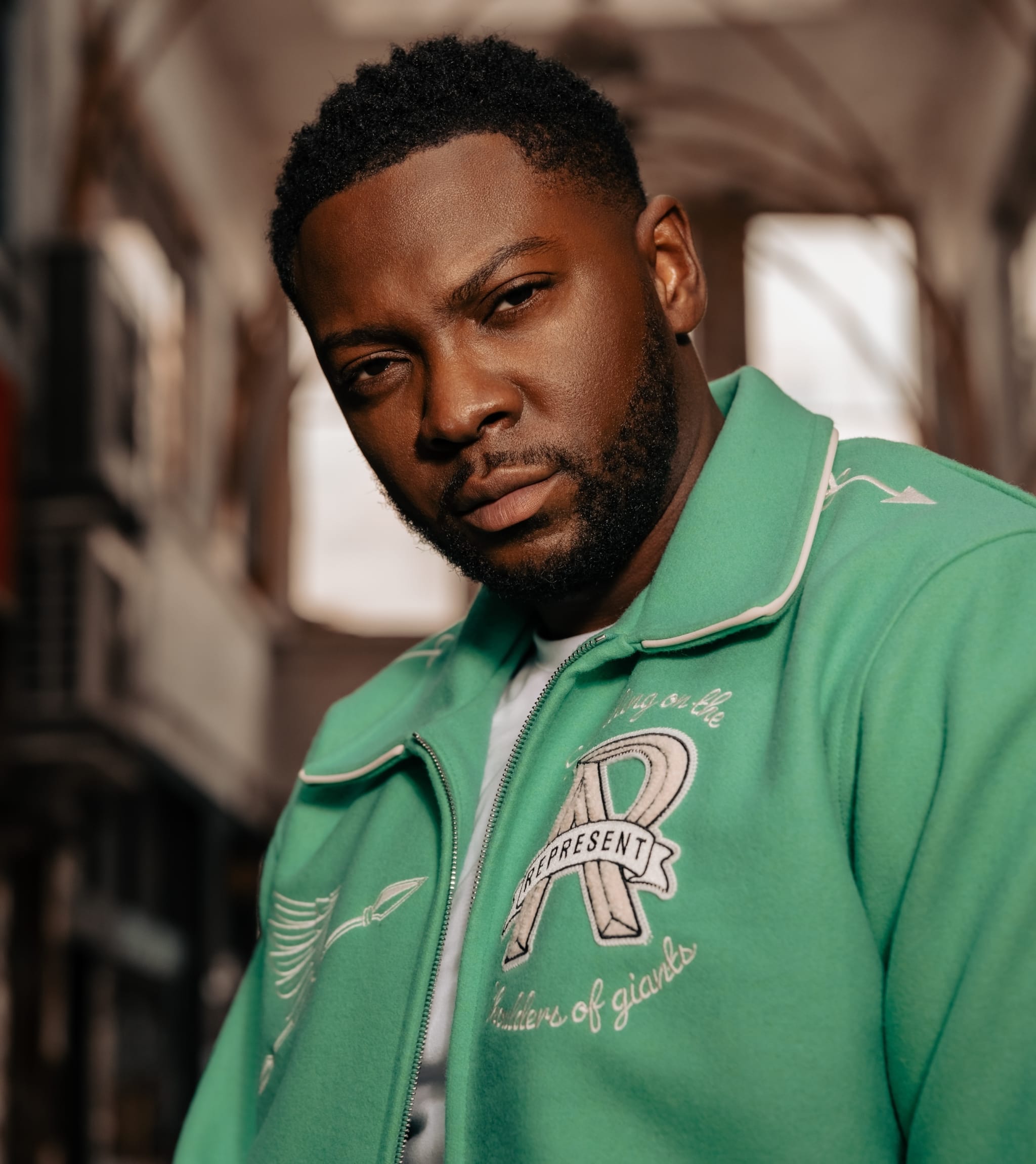 A man with a serious expression is wearing a green jacket with detailed embroidery, standing in an urban setting