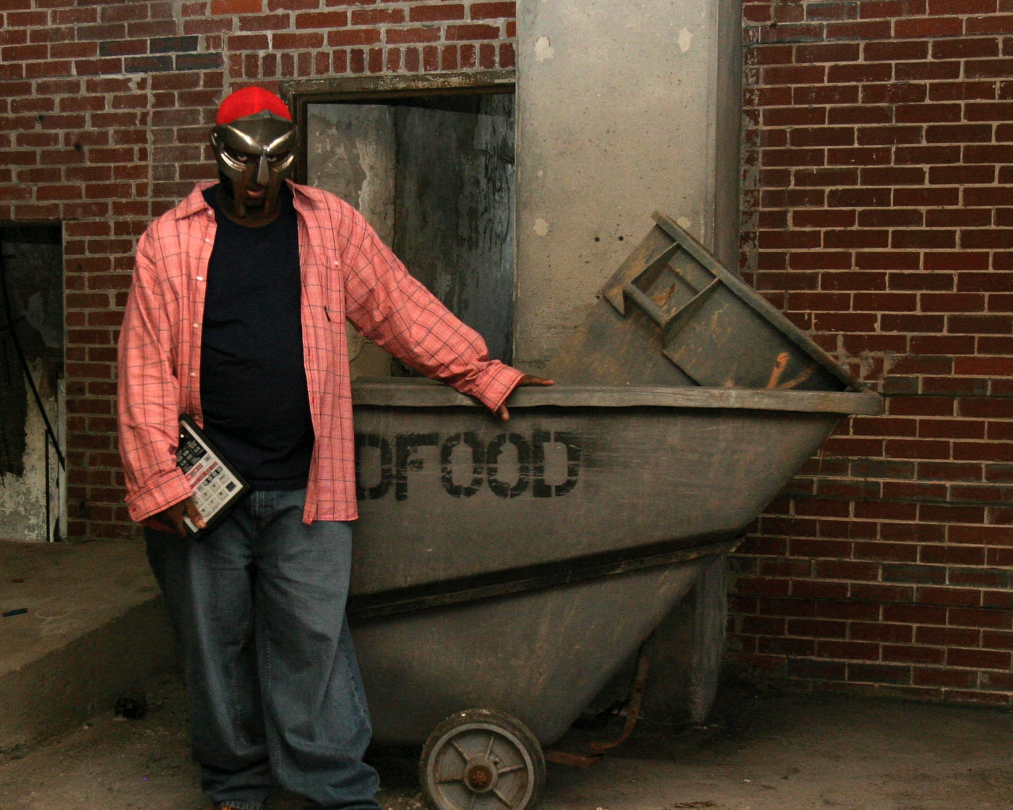 MF DOOM stands by a brick wall, holding a device, next to a large industrial container.