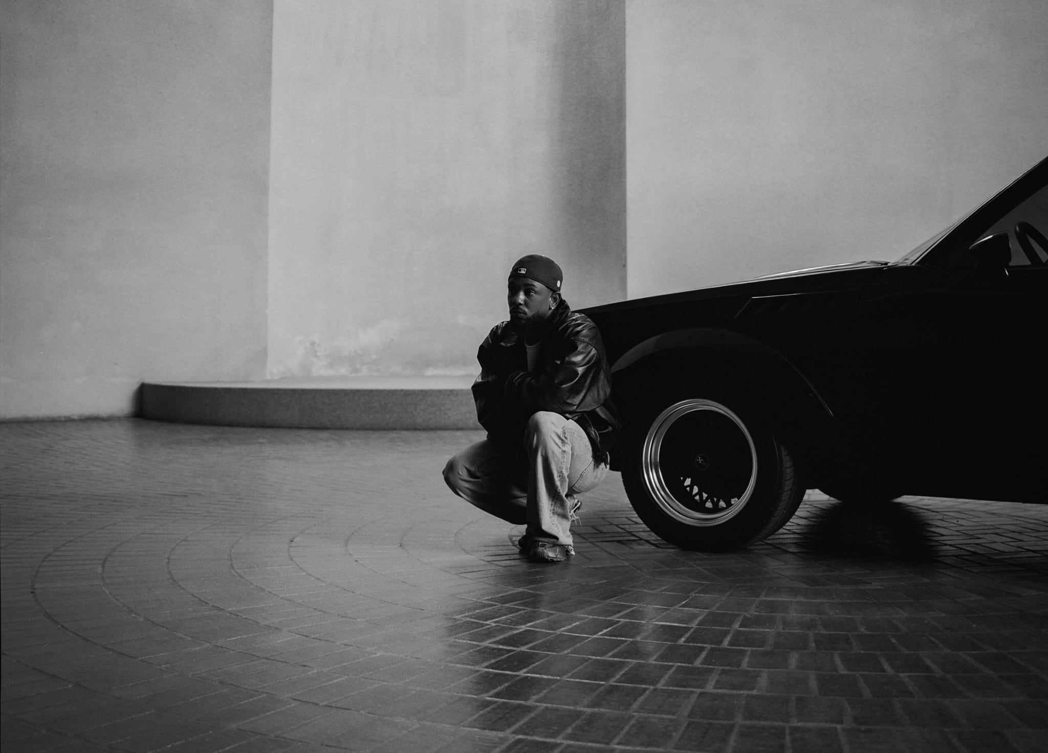Kendrick Lamar squats beside a black car on a tiled floor, wearing a leather jacket and cap, in a minimalist setting.