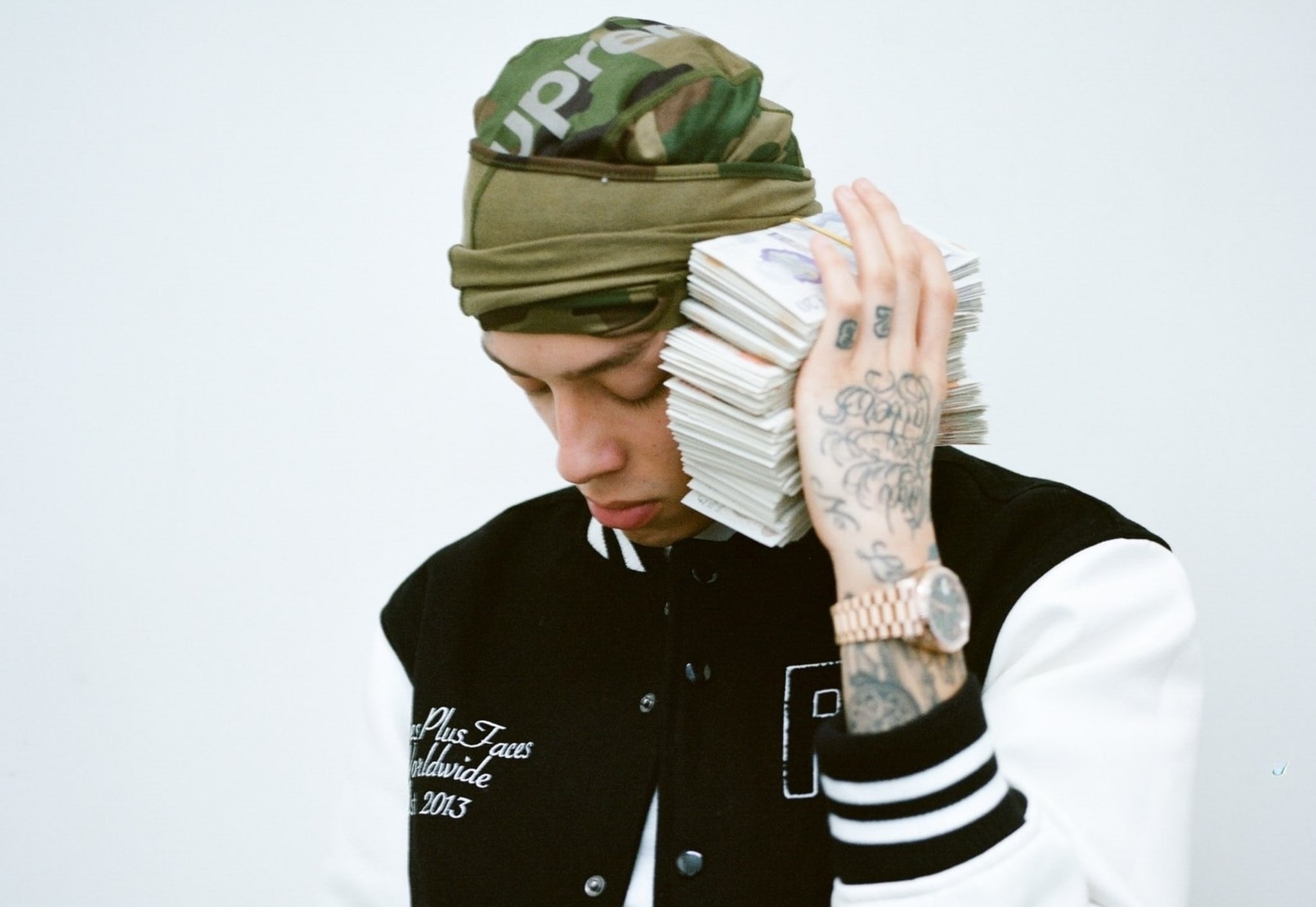 A person wearing a camo head wrap and a varsity jacket holds a large stack of cash to their ear like a phone. Tattoos are visible on their hand and neck