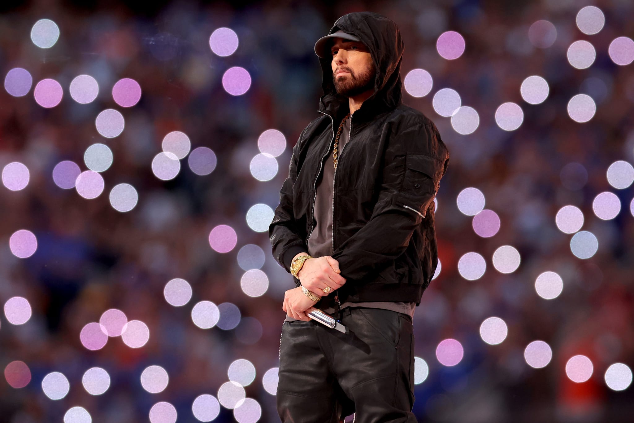 Eminem performing on stage, wearing a black hoodie and jacket, holding a microphone. The background is filled with blurred lights.