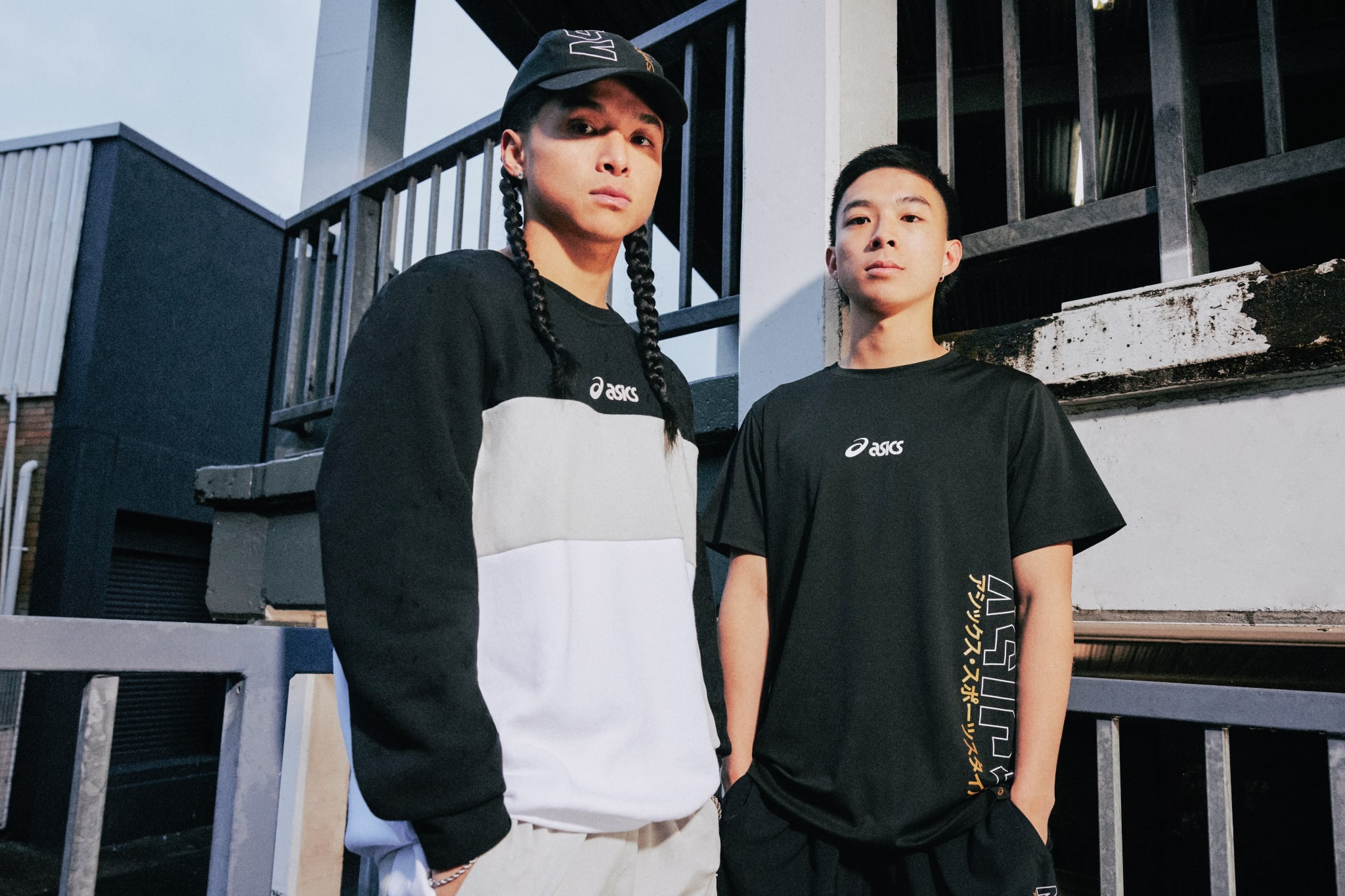 Two young men wearing Asics clothing stand outdoors. One has braided hair and a cap, and the other has short hair. They pose confidently.