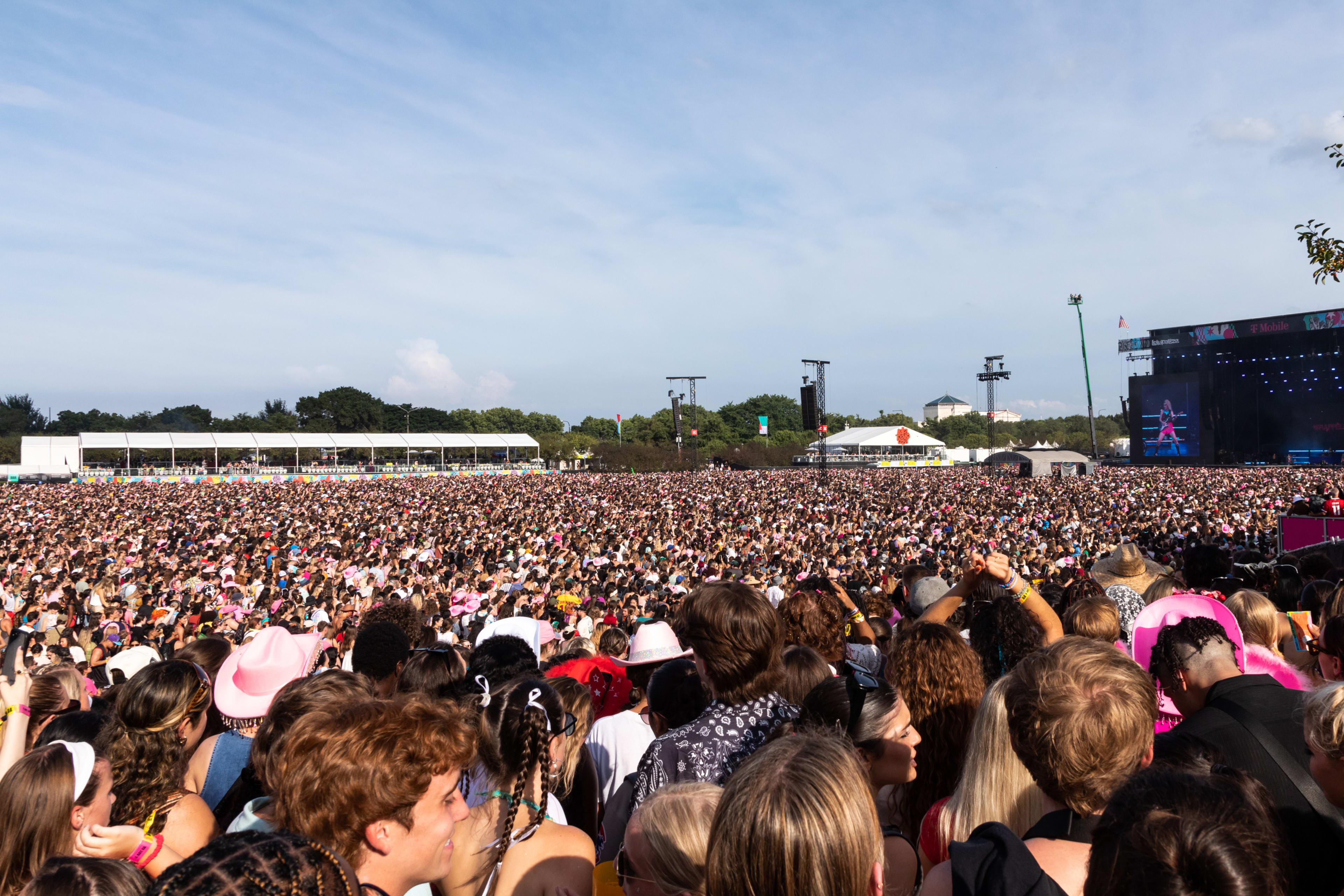 Large crowd at an outdoor music festival, facing a distant stage, with some attendees in festival attire. Musicians are not visible