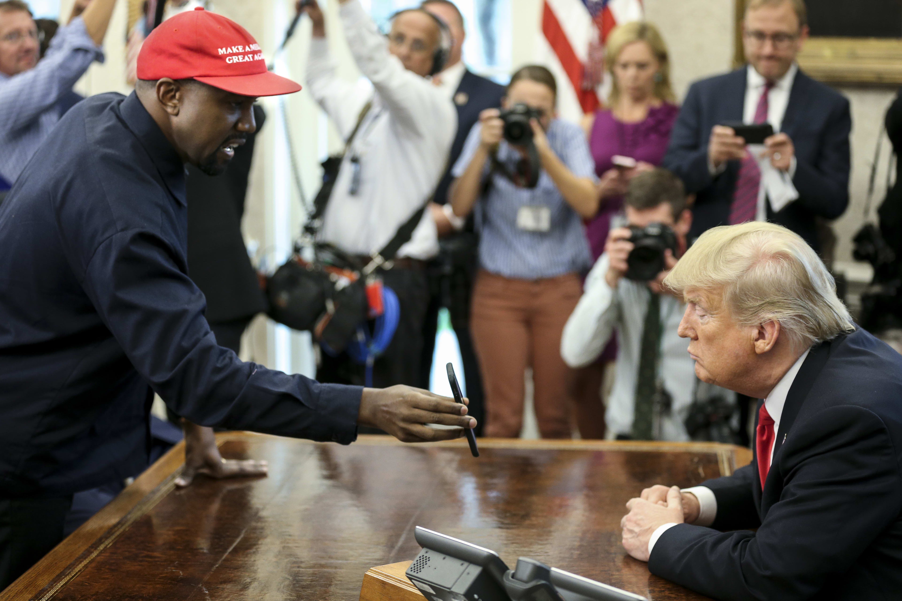 Kanye West, wearing a "Make America Great Again" cap, hands a pen to Donald Trump, who is seated at a desk surrounded by media, in what appears to be an official setting