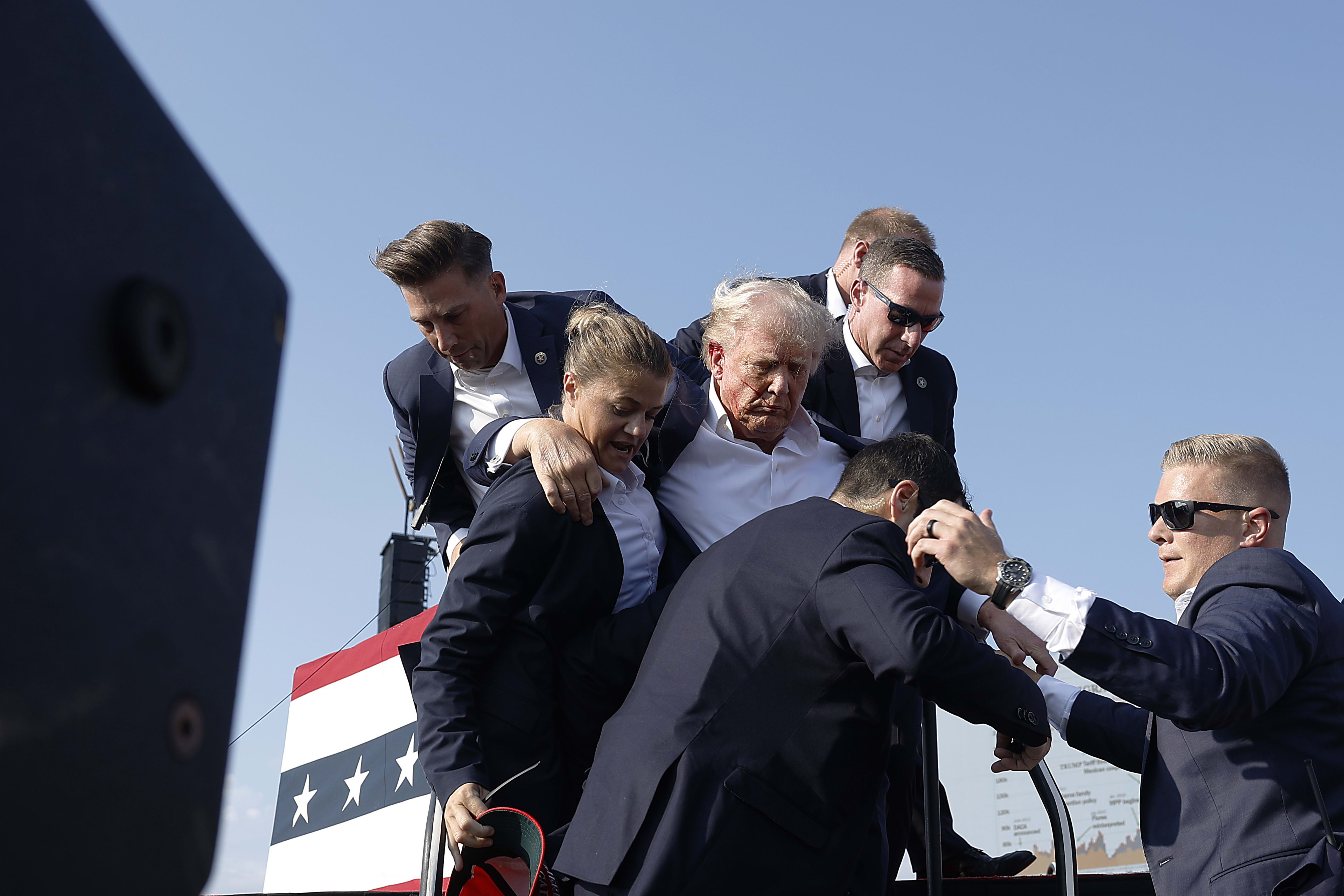 Donald Trump, surrounded by security personnel, being assisted by five men, on a stage with a red, white, and blue backdrop