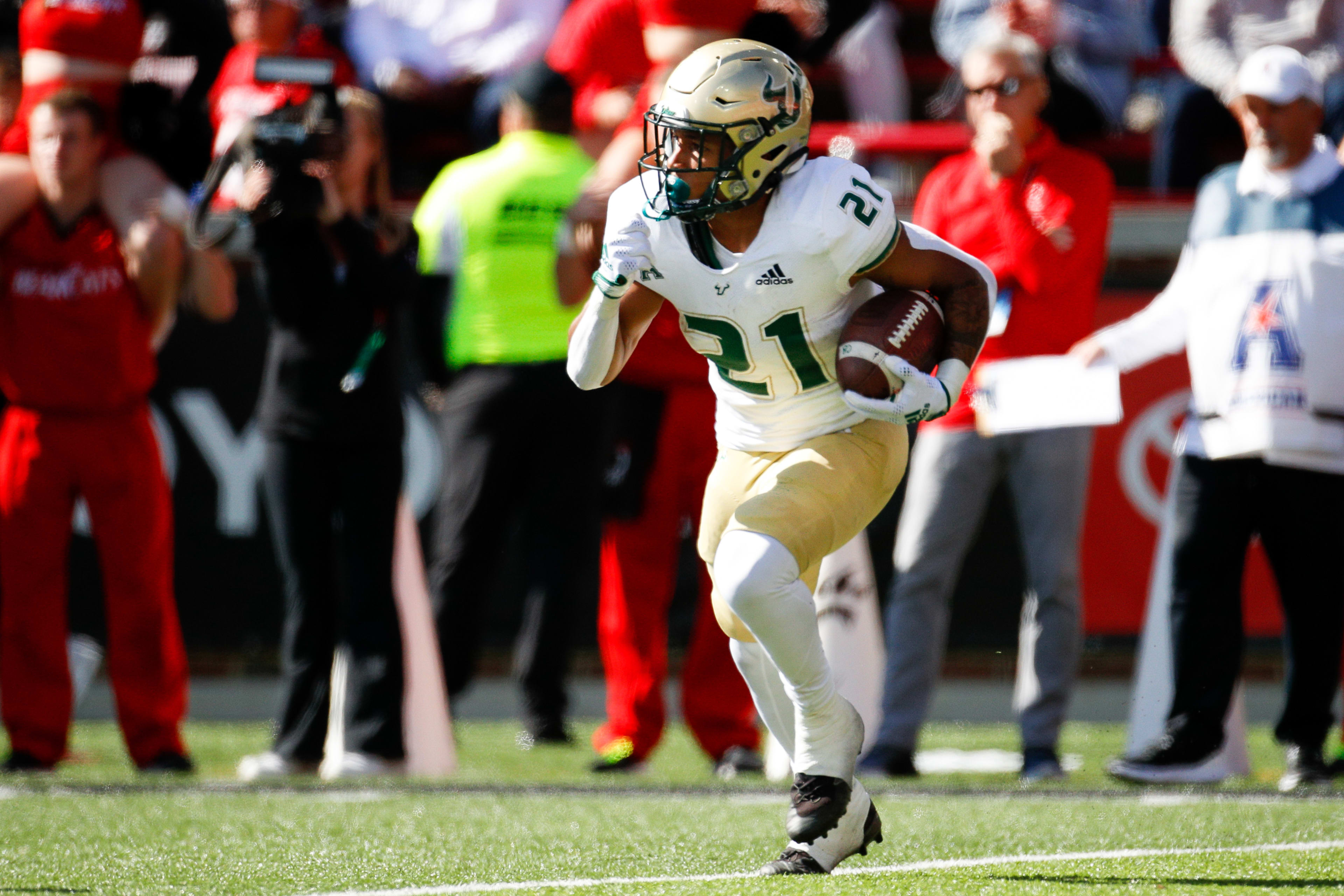 A football player, number 21, runs with the ball during a game. Spectators and photographers are seen in the background