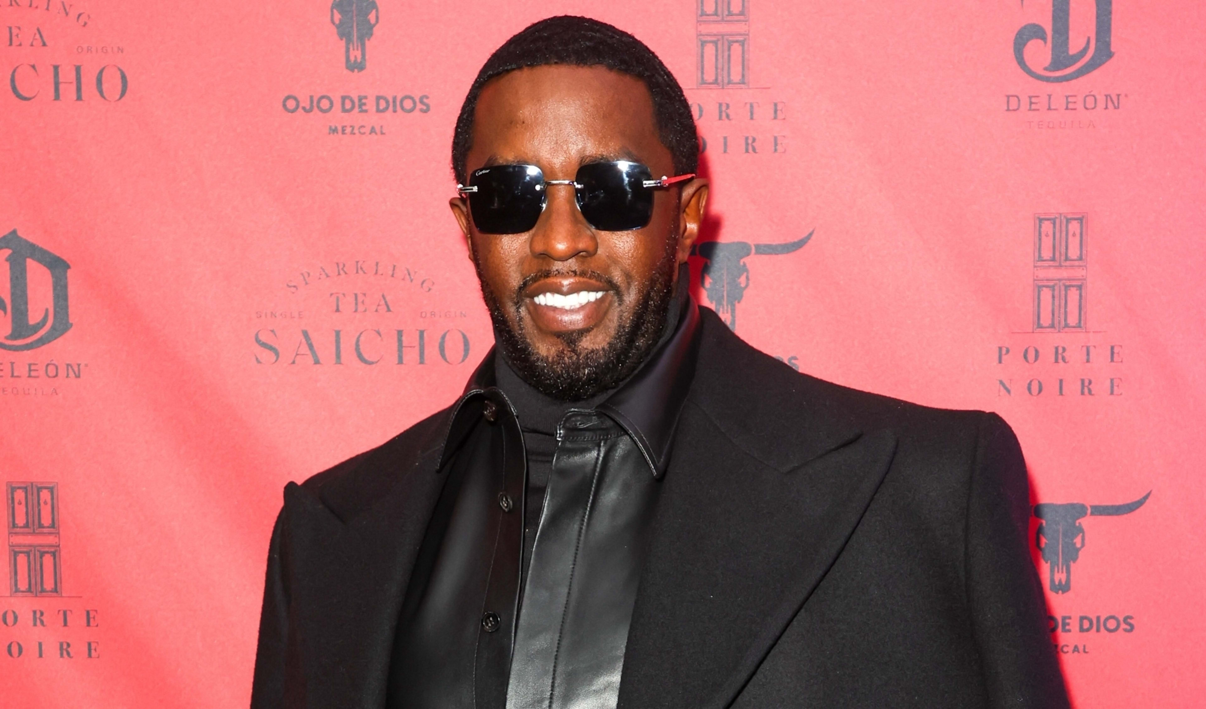 Man in sunglasses and black outfit at event, posing for cameras on brand background
