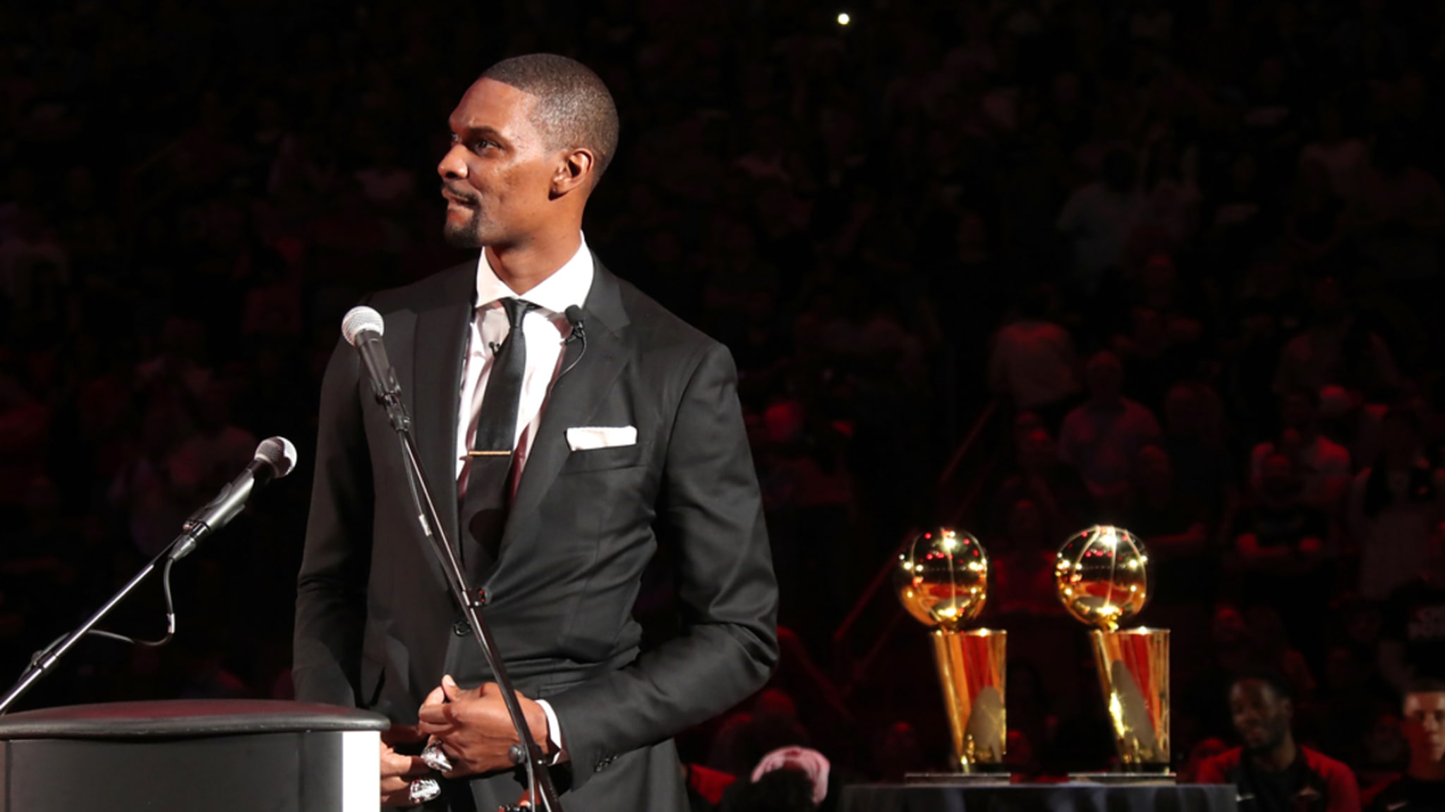 Chris Bosh stands at a podium in a suit during an event. Two NBA championship trophies are displayed on a table beside him