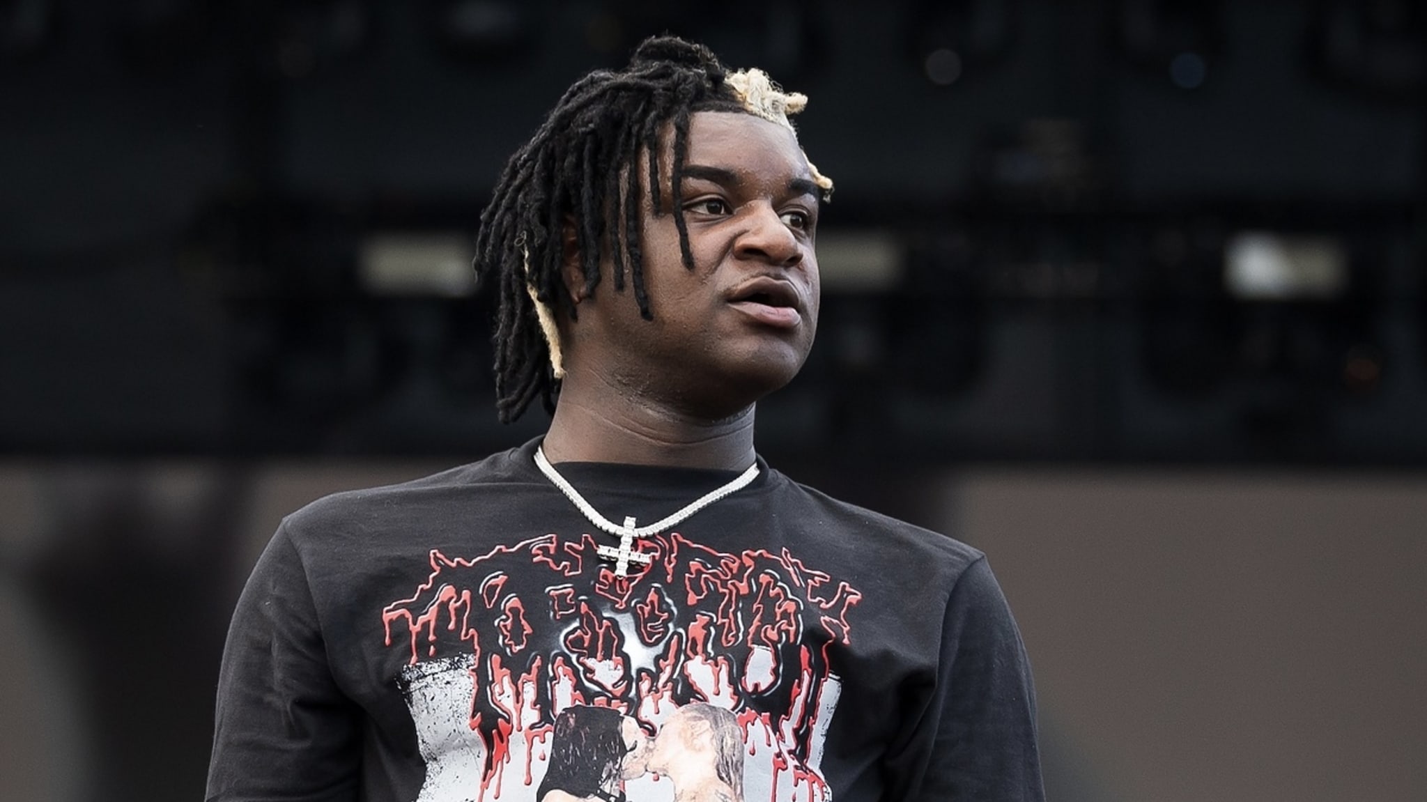 Musical artist facing forward, wearing a graphic t-shirt with an edgy design, layered necklace, and distinctive hairstyle