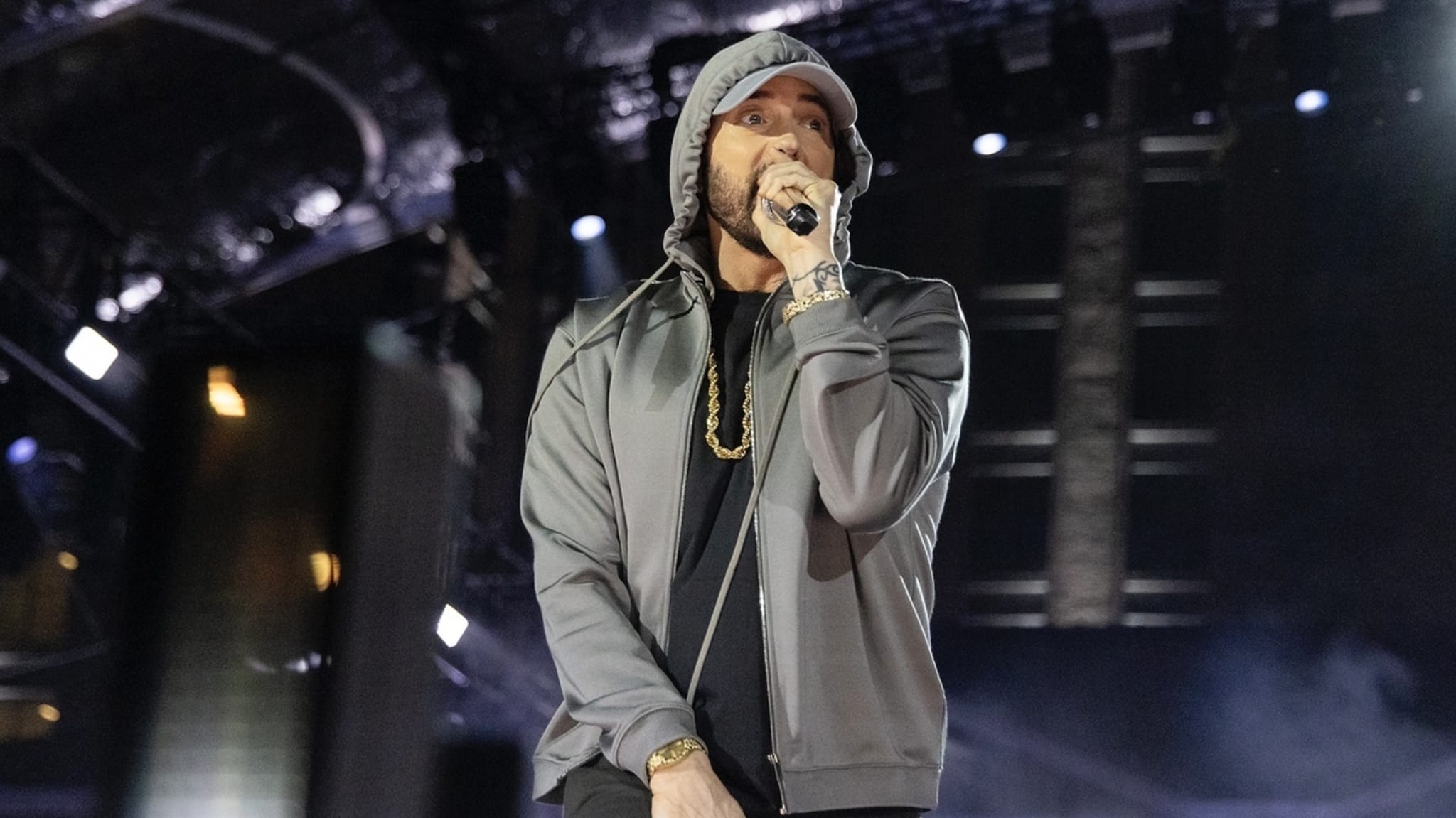 Eminem performs on stage wearing a hoodie and chain necklace, holding a microphone