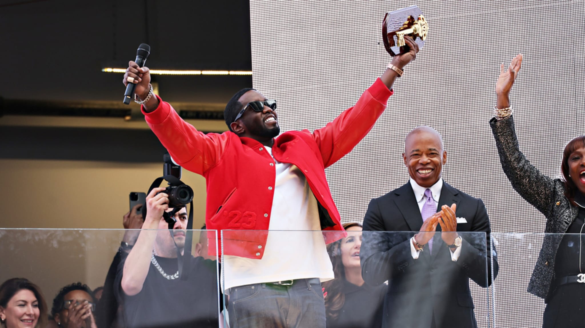 Person in a red jacket holds an award and cheers on stage. Another person in a suit stands next to him, applauding. People in the audience also cheer and clap