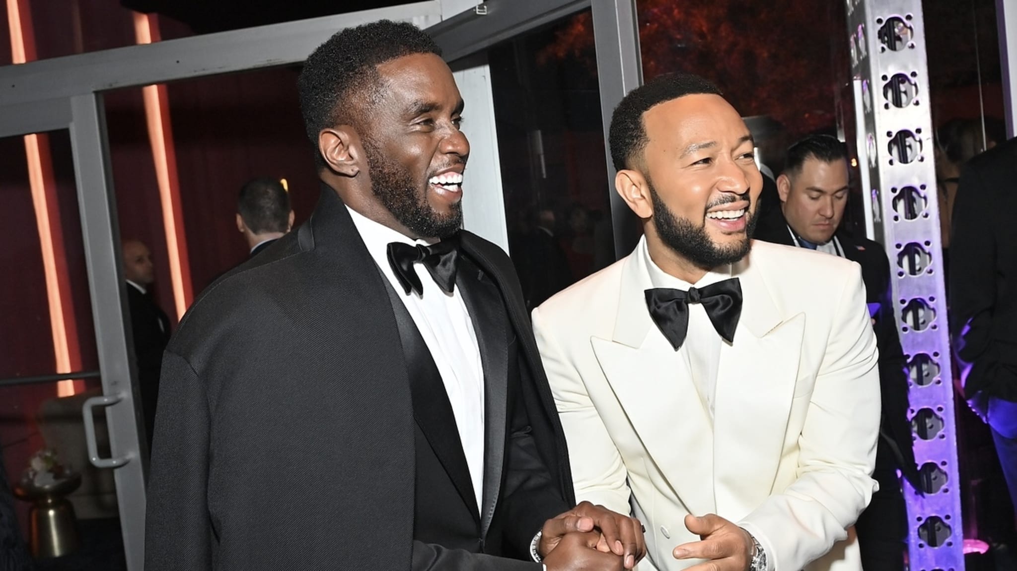 Sean Combs and John Legend are smiling and shaking hands at a formal event. Sean is wearing a black suit with a cape, and John is in a white tuxedo