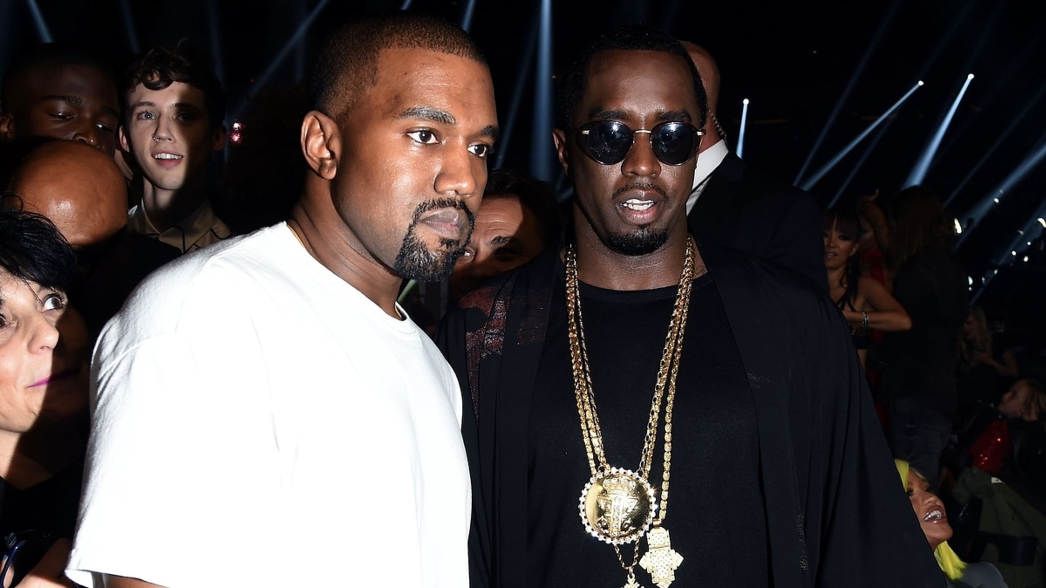 Two men at a music event, both dressed in dark outfits with prominent gold jewelry