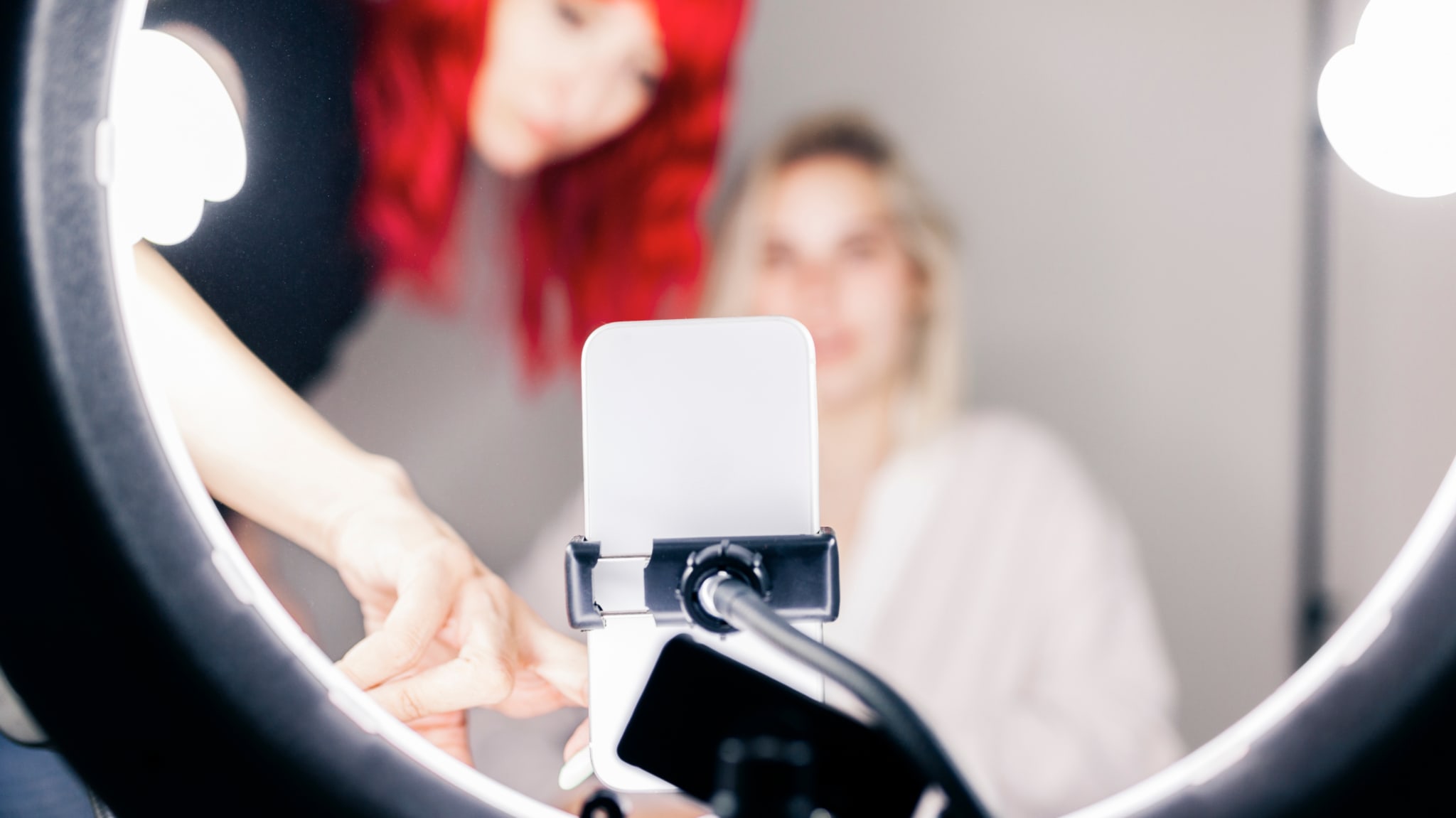 Two influencers are seen through a ring light, setting up a smartphone for recording or photography. One has vibrant red hair.