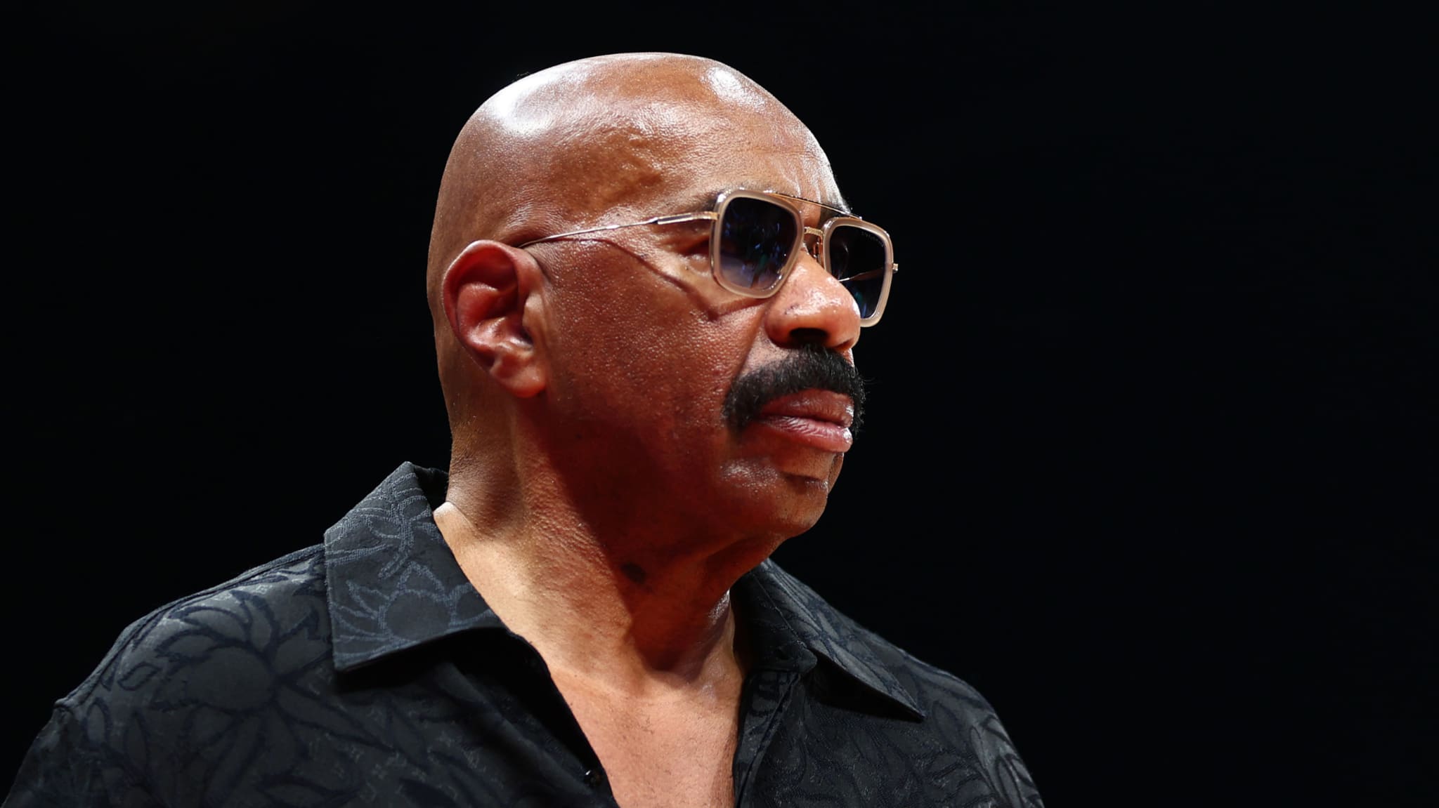 Steve Harvey wearing sunglasses and a patterned black shirt, looking to the side.