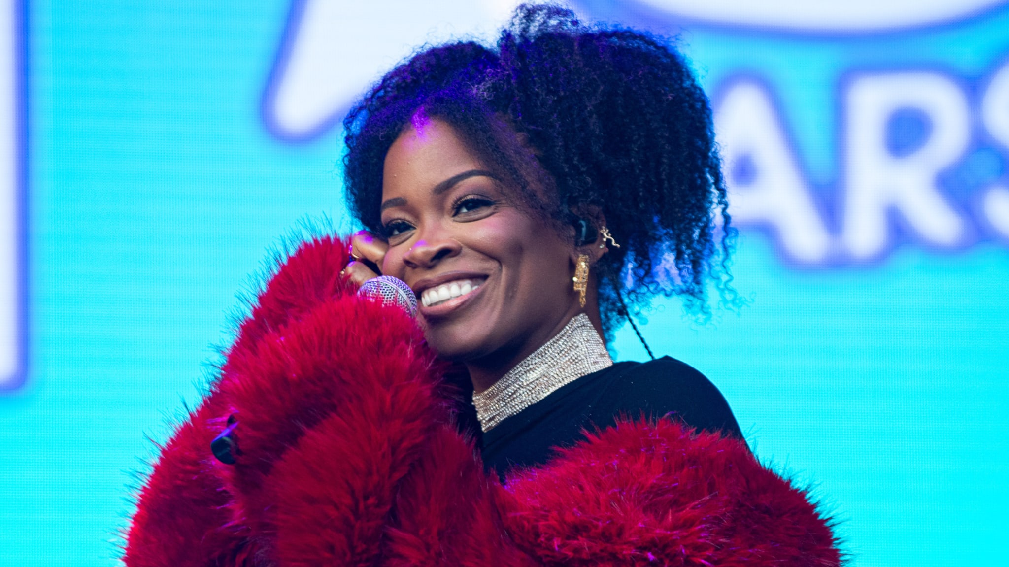 A woman with curly hair, wearing a red fur coat, smiles while holding a microphone on stage.