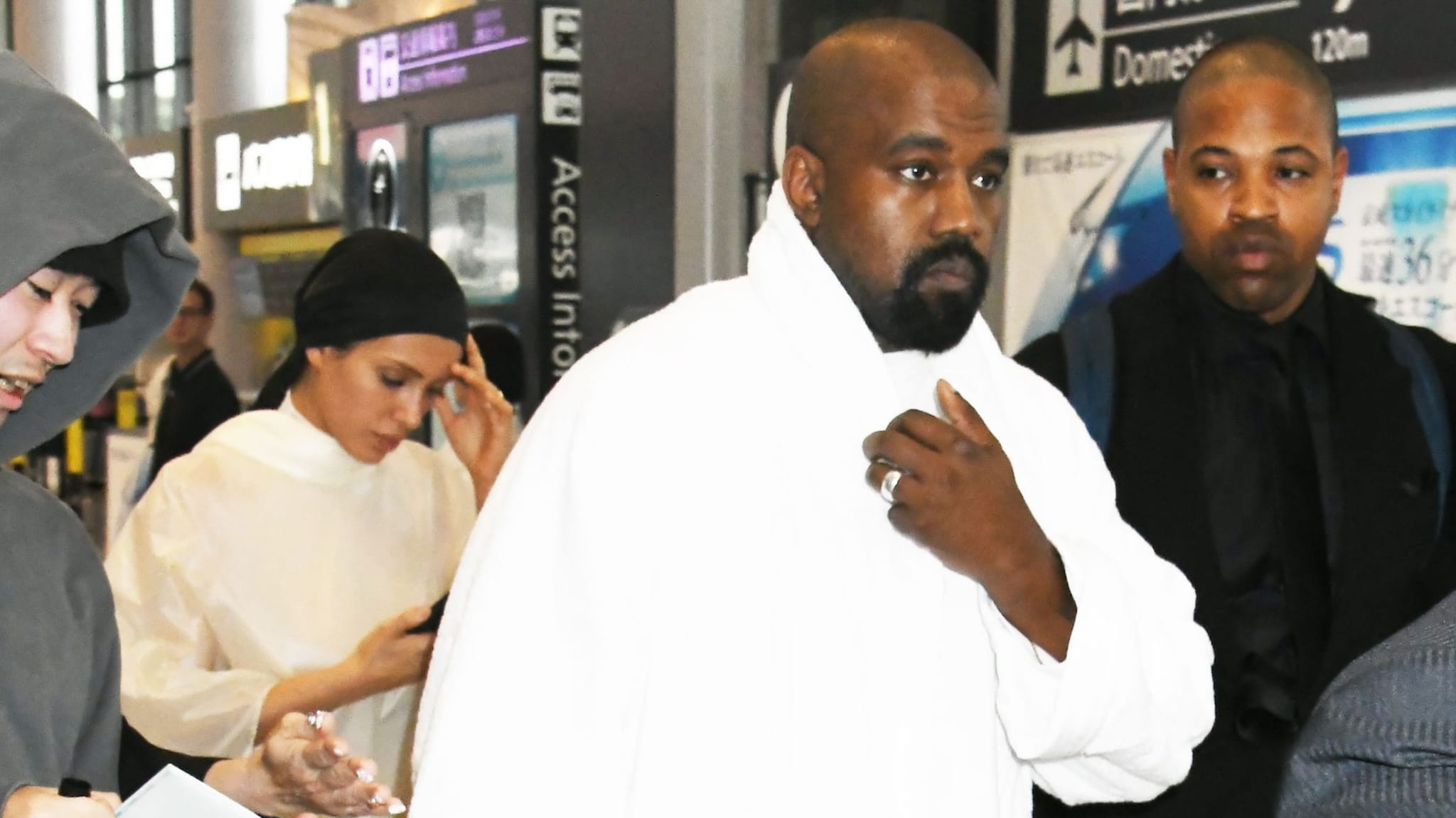 Kanye West wearing a white outfit at an airport, surrounded by people.