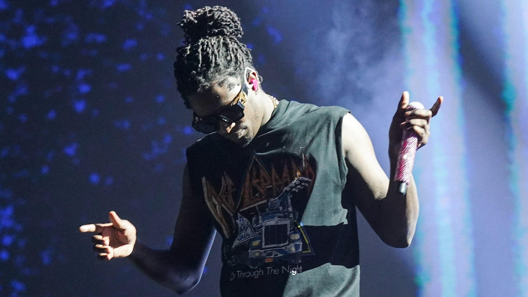 Young Thug on stage with sunglasses and a Def Leppard shirt, holding a microphone, with a spotlight in the background.