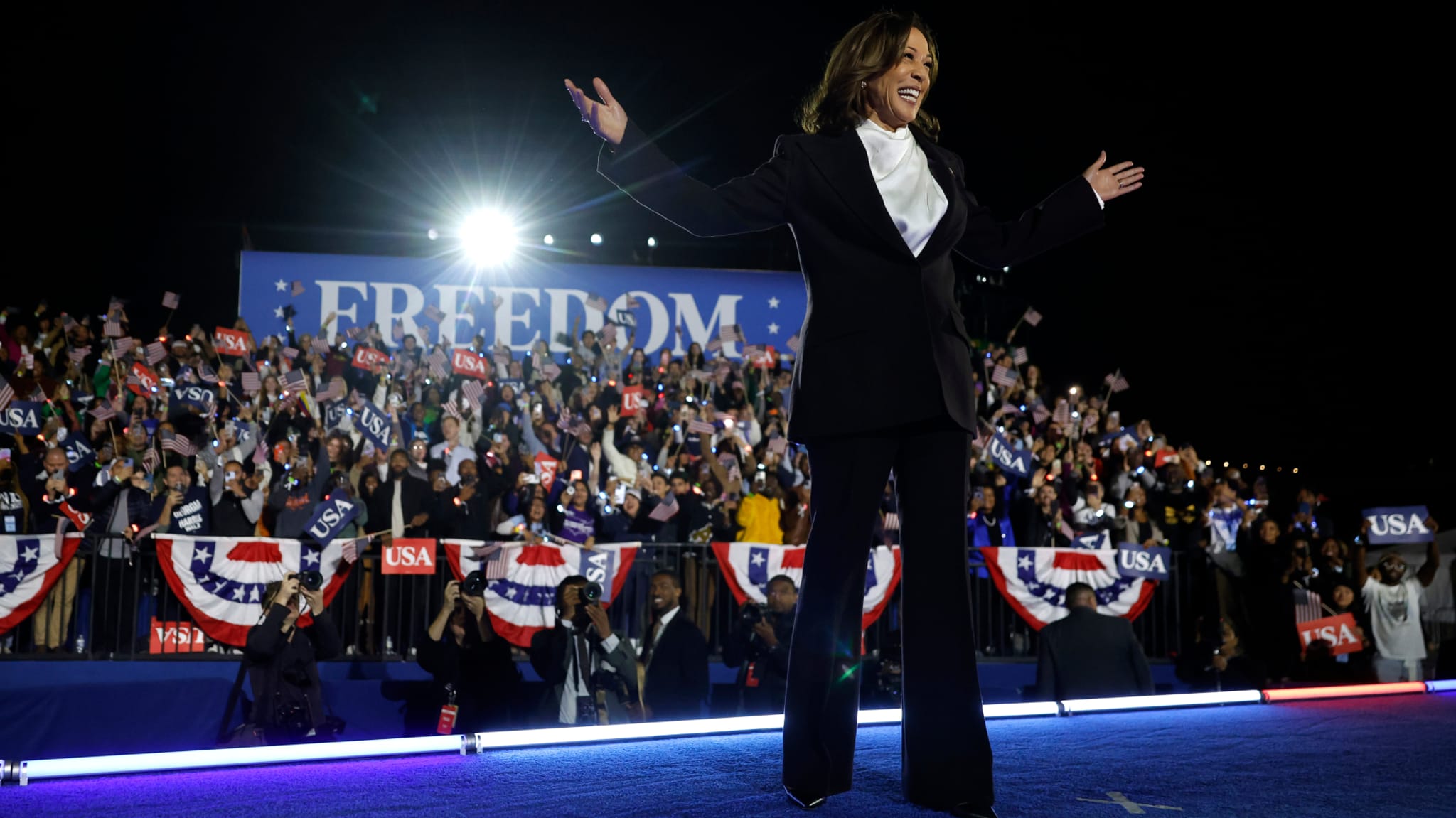 Kamala Harris on stage, arms raised, in front of a cheering crowd with "Freedom" sign and USA flags.