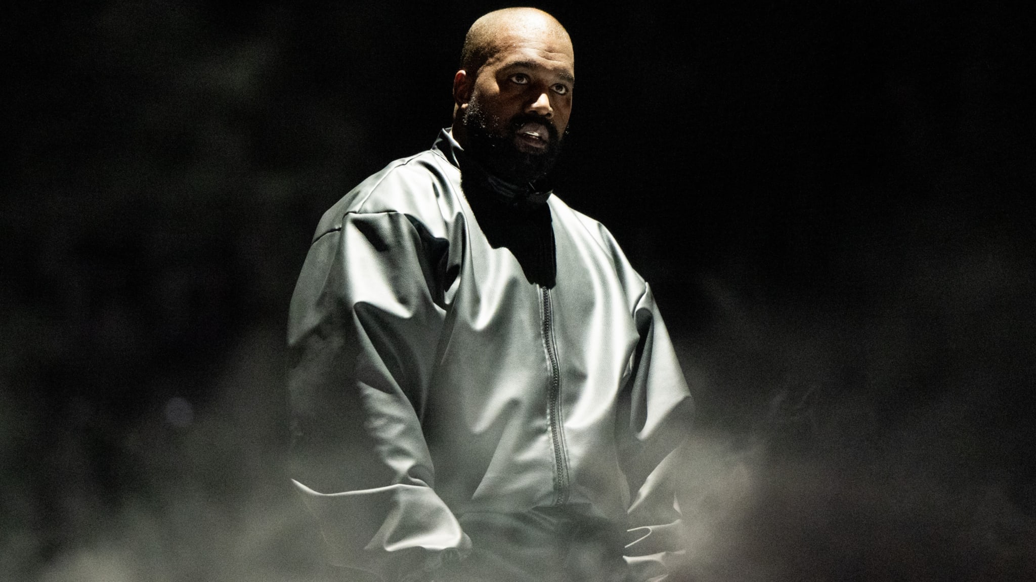 Ye in a dark, oversized jacket, standing amidst a foggy, dimly lit background.