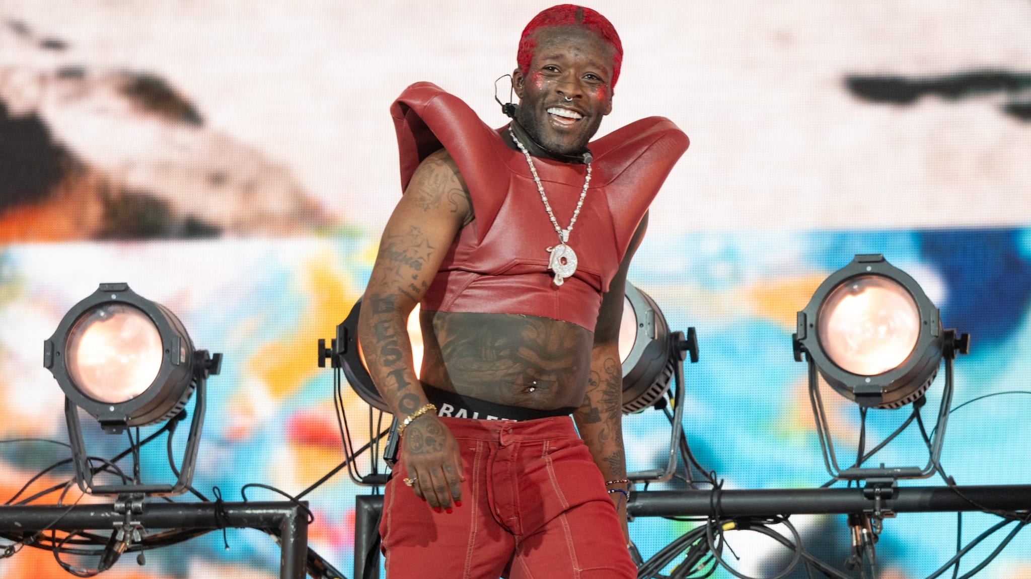 Lil Uzi Vert performing on stage, wearing a red outfit with shoulder pads and a necklace, smiling with stage lights in the background.