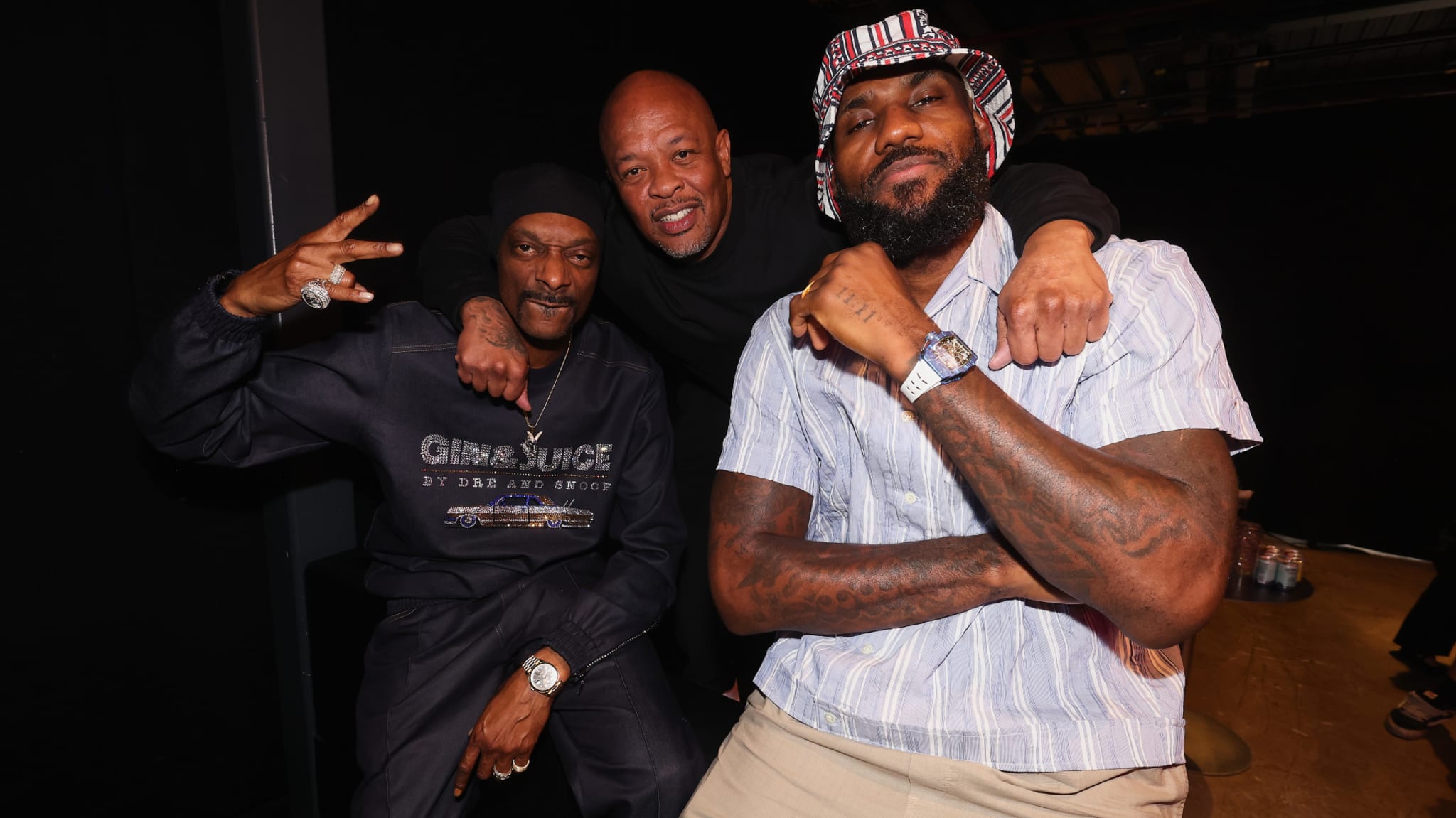 Snoop Dogg, Dr. Dre, and LeBron James posing together. Snoop flashes a peace sign, Dr. Dre smiles, and LeBron crosses his arms.