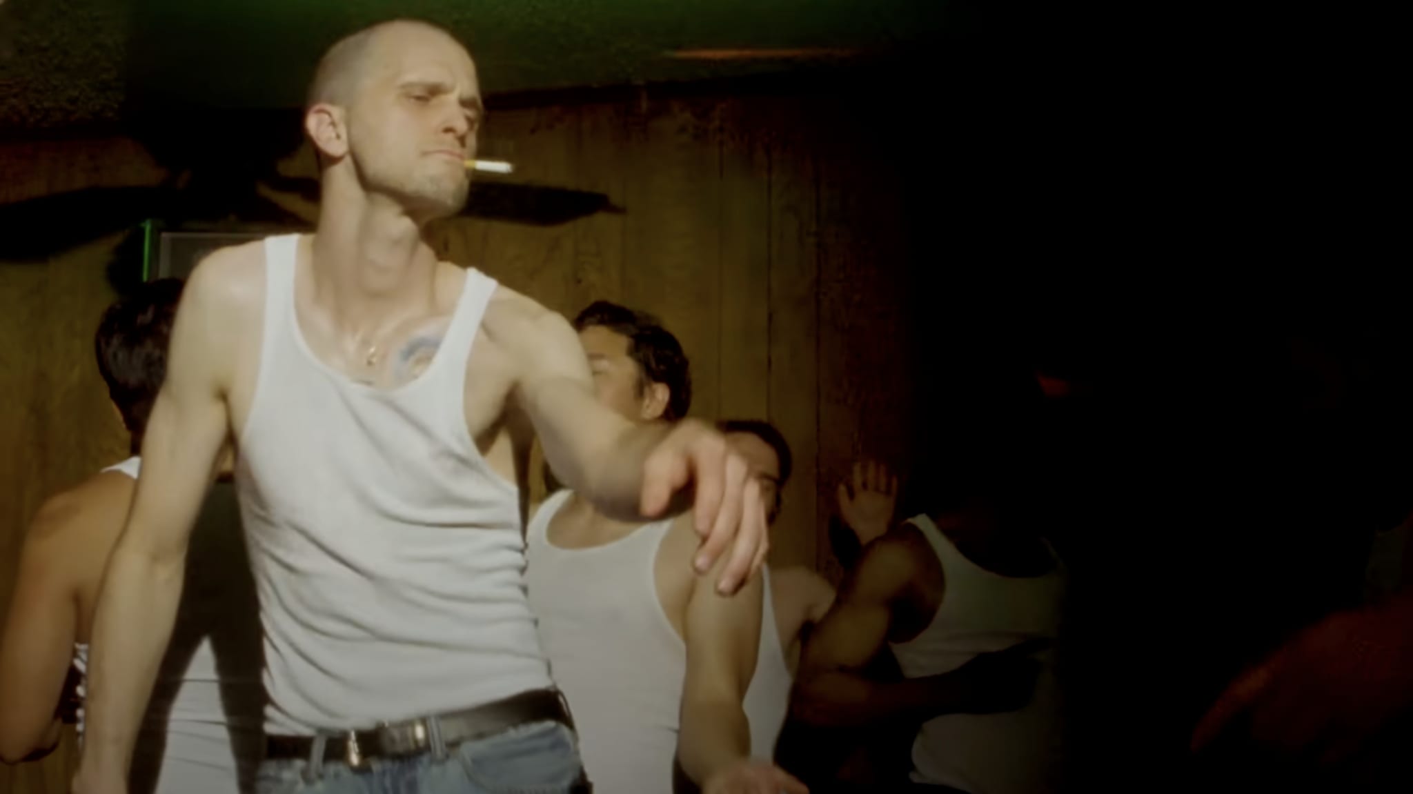 JMSN, who is wearing a white tank top and jeans, dances with a cigarette in his mouth, surrounded by others in a dimly lit room.