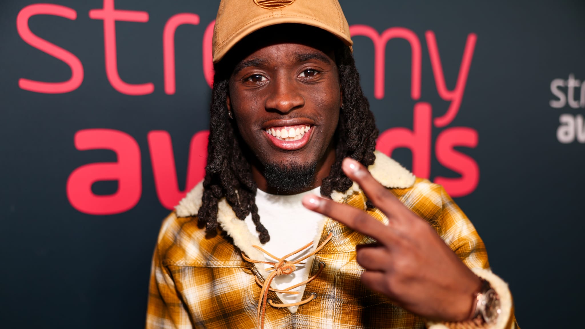 Man smiling, wearing a yellow plaid jacket and a tan cap, making a peace sign at the Streamy Awards backdrop.