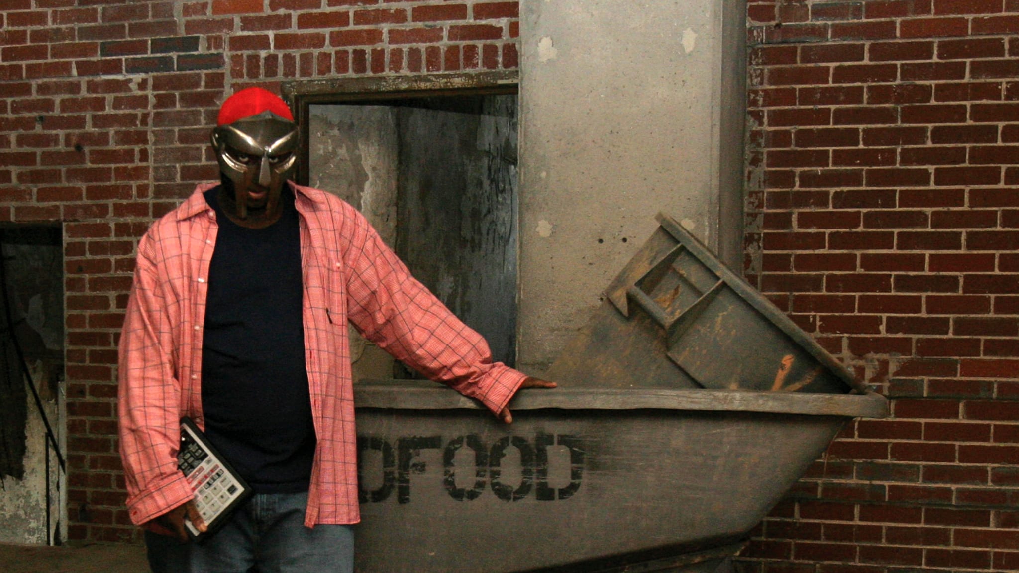 MF DOOM stands beside a large metal container in an industrial setting.