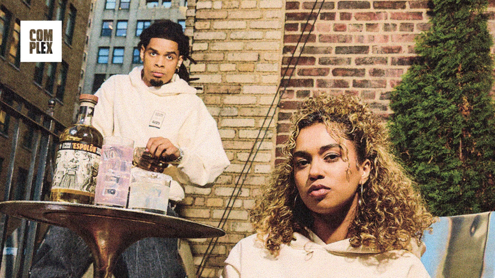 Two people in casual outfits, one sitting cross-legged in the foreground, the other standing by a table with bottles, against a brick wall.