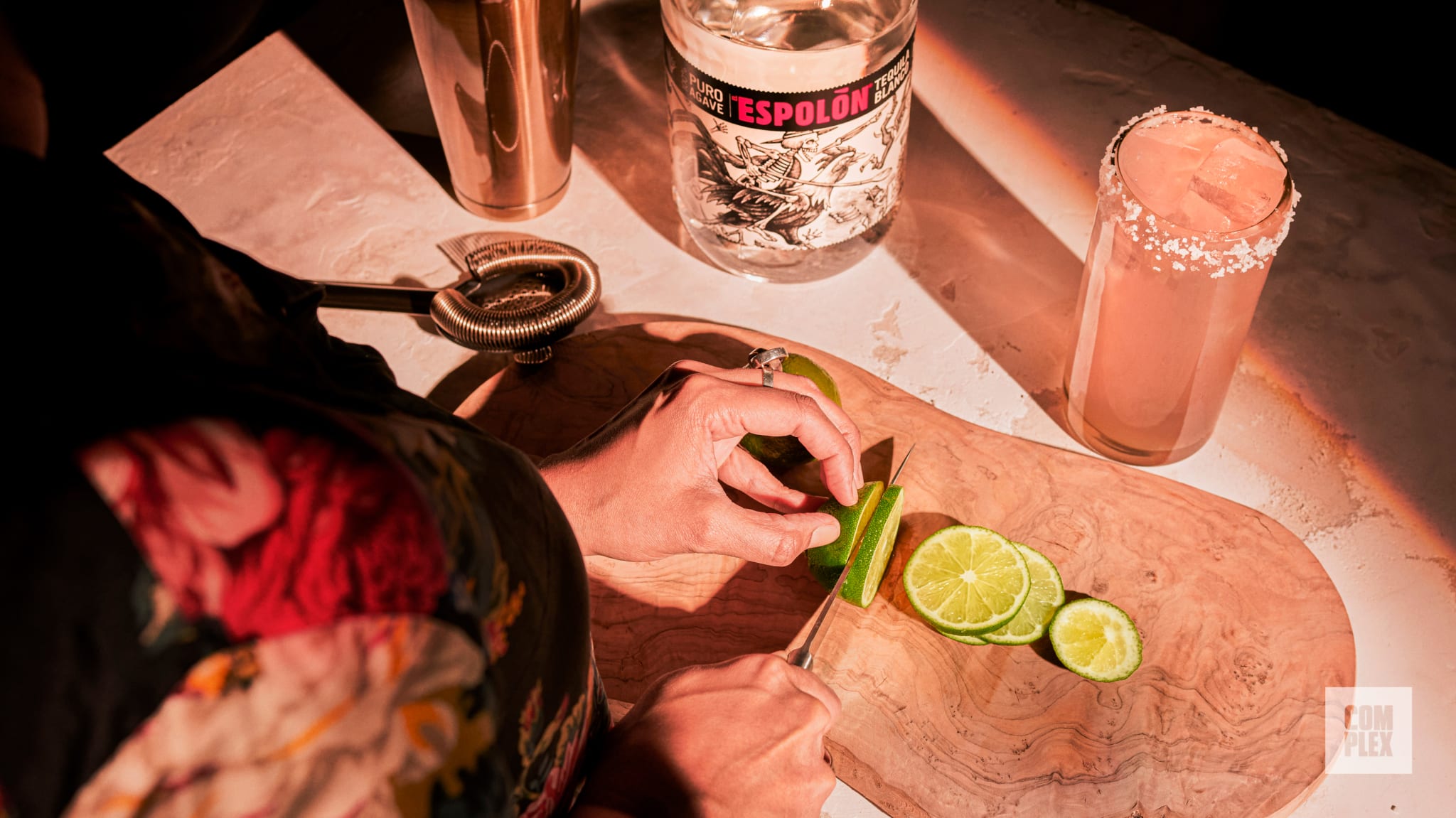 A person slices limes on a wooden board. Nearby are a bottle of Espolón tequila, a cocktail shaker, and a salt-rimmed drink.