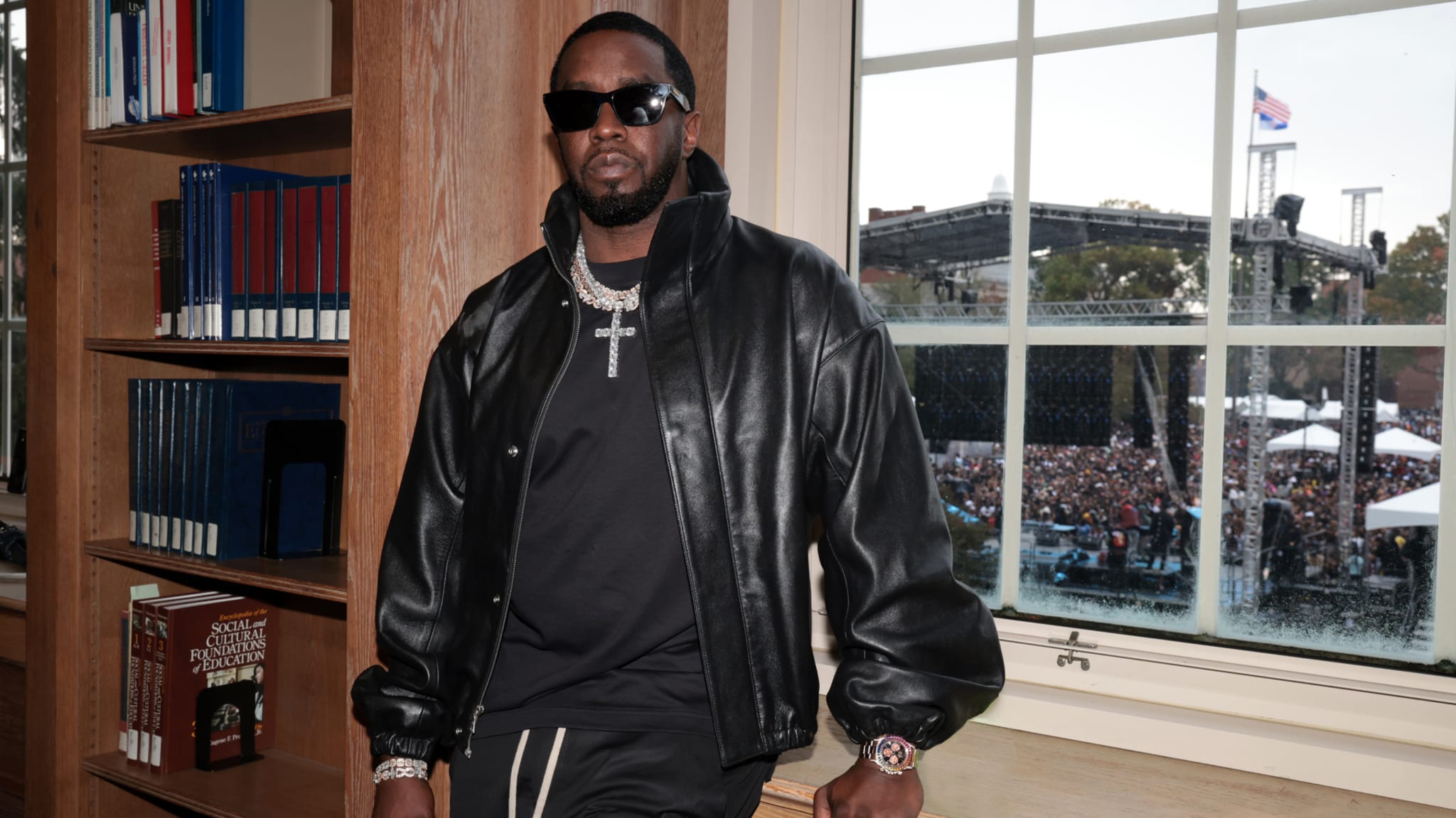 Sean "Diddy" Combs in a black leather jacket and sunglasses, standing by a window with a crowd and stage visible outside.