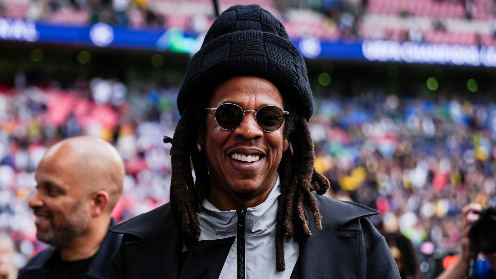 Jay-Z smiling, wearing sunglasses and a black hat, at a sports event with a crowd in the background.