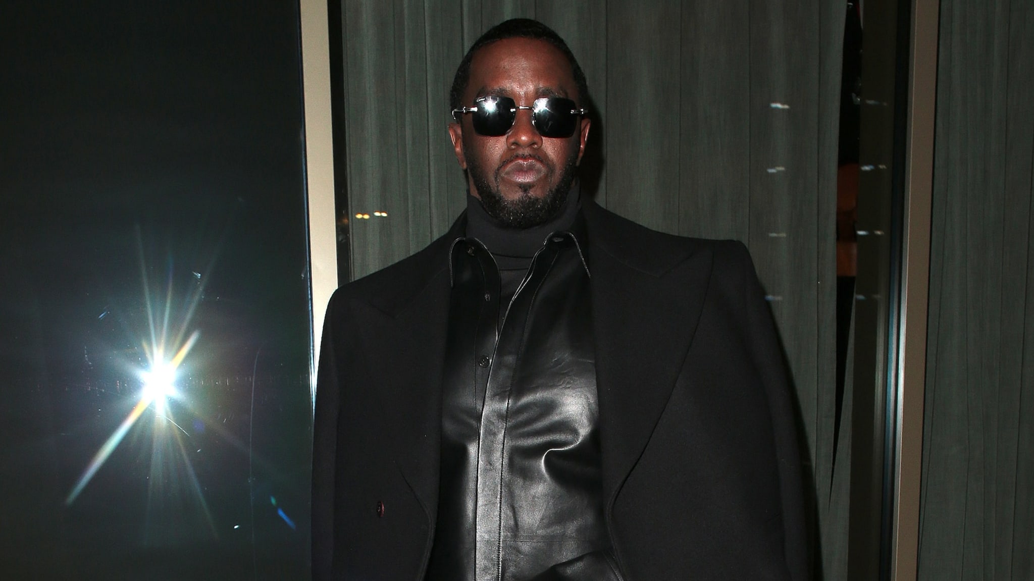 Sean "Diddy" Combs in a black leather outfit and sunglasses, standing indoors.