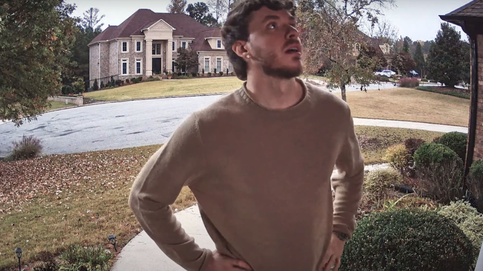 Jack Harlow in a brown sweater stands on a porch, looking up. A large house and trees are visible in the background.