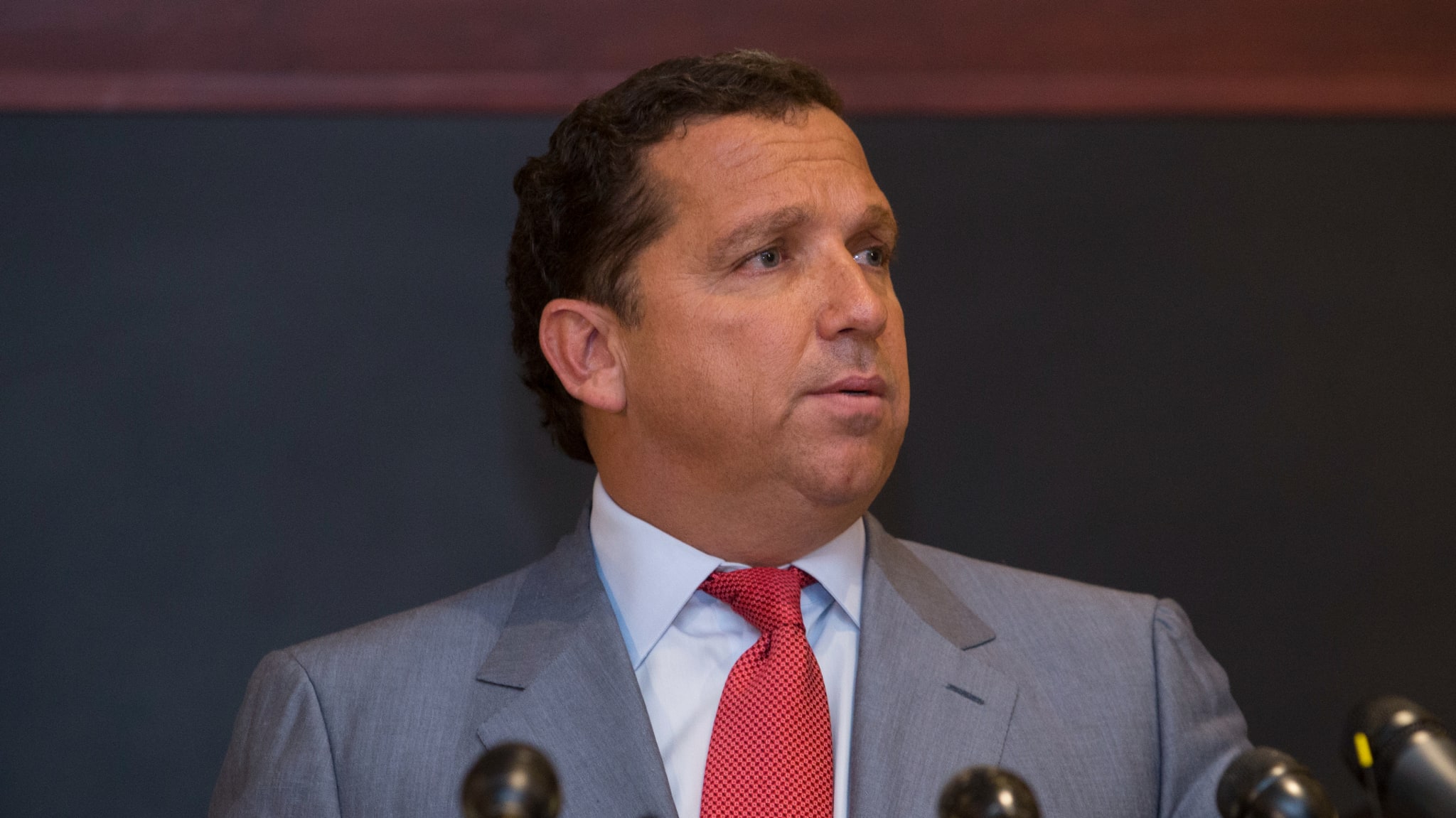 A man in a gray suit and red tie speaks at a podium with microphones.