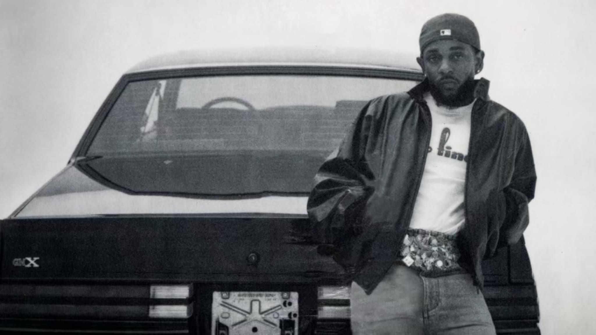 Kendrick in a leather jacket and cap leans against a vintage car, with a relaxed pose and hands in pockets.